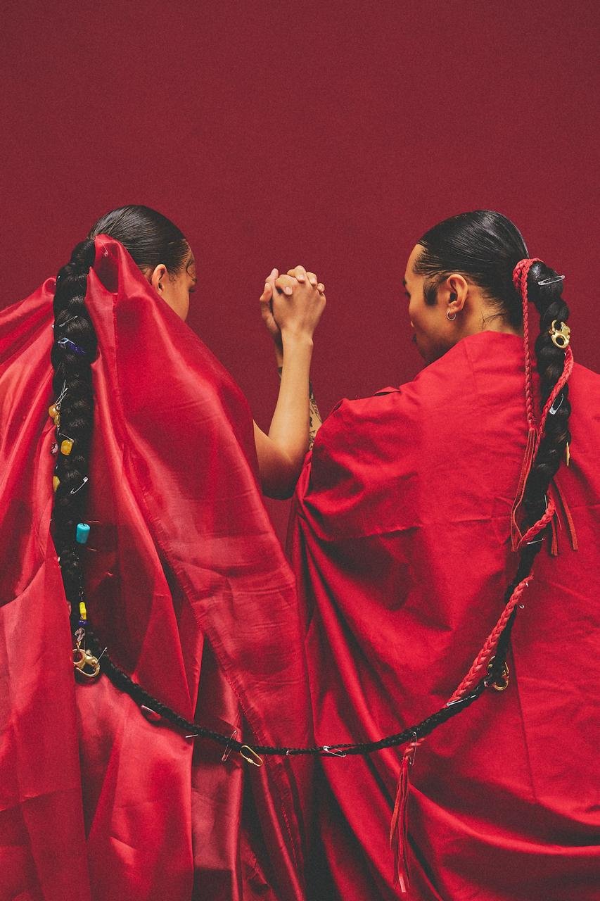 Deux femmes avec des cheveux noirs tressés, drapées dans des tissus rouges, se tiennent la main lors d'une épreuve. Composition artistique avec fond rouge.