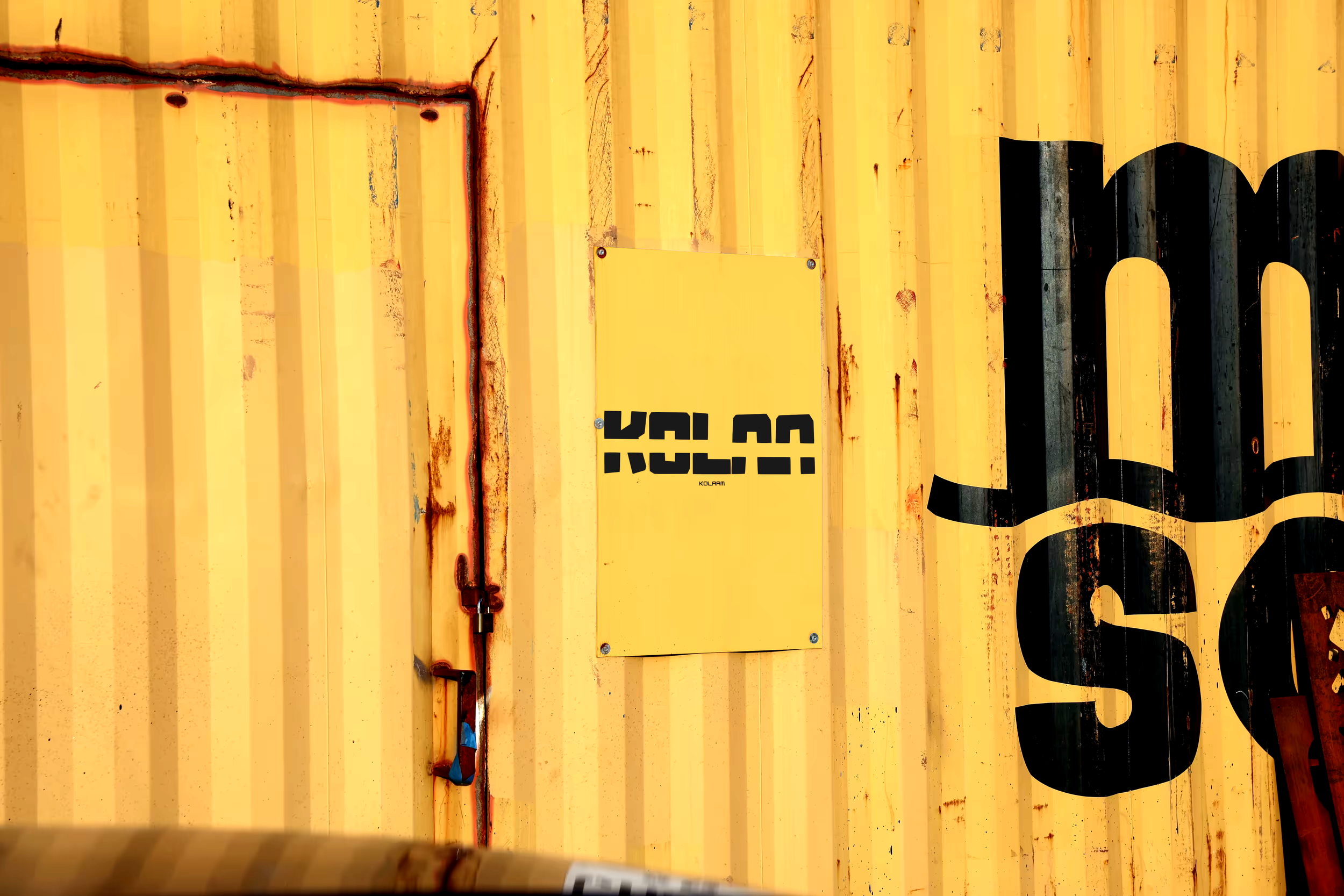 Portière en métal jaune avec une plaque jaune portant le mot 'KOLAS' en noir, montrant des signes de rouille et un cadenas.