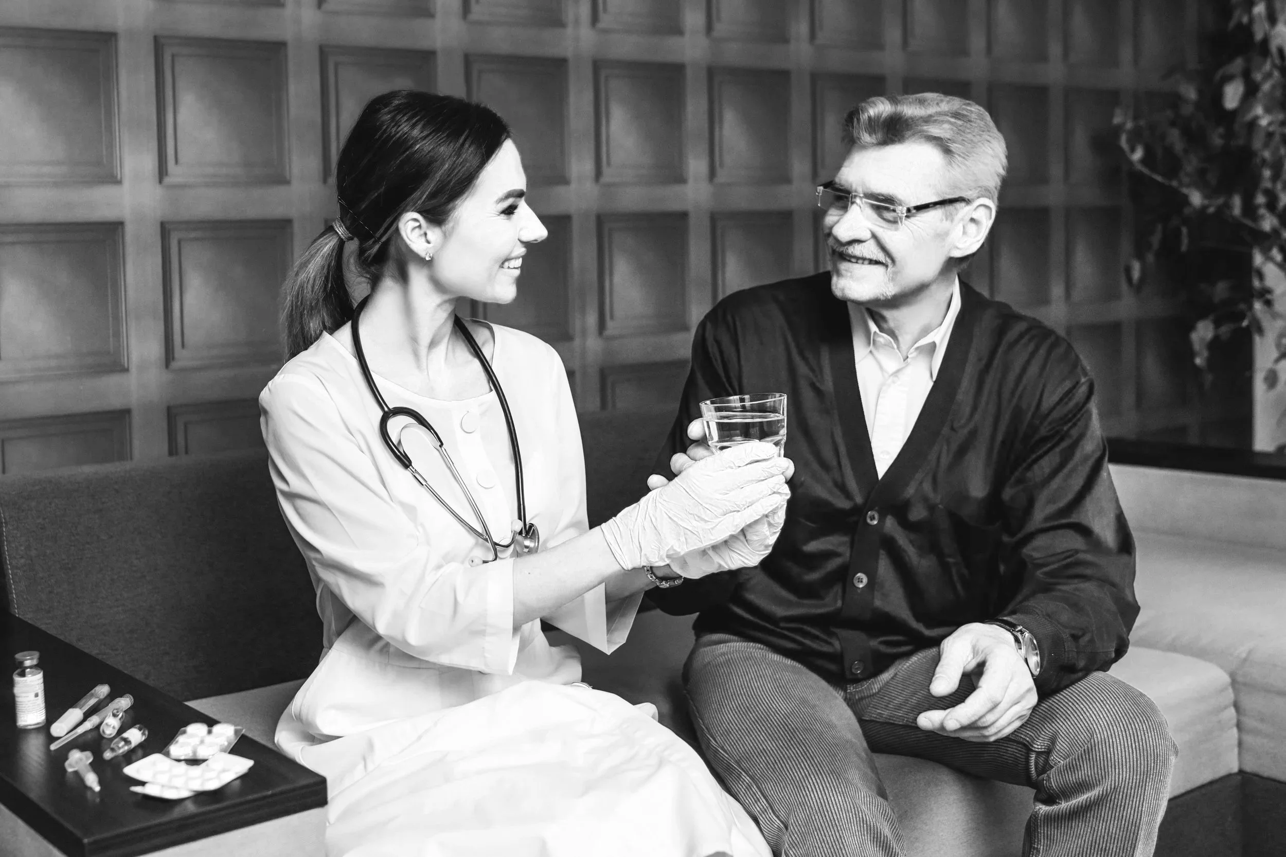 A female healthcare worker in a white coat and stethoscope, wearing gloves, handing a glass of water to a smiling older man sitting on a couch in a room with wood-paneled walls.