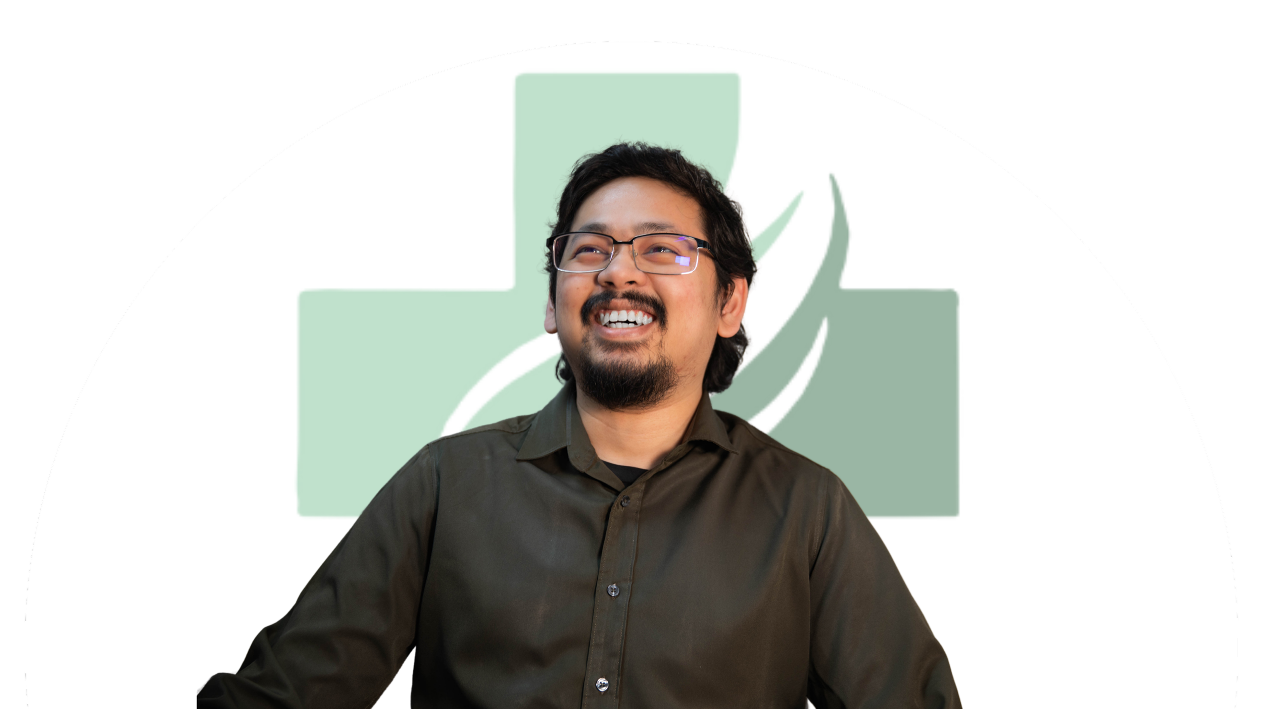 A man with glasses, dark hair, and a goatee, smiling and looking slightly upward, standing in front of a green medical cross with a leaf design.