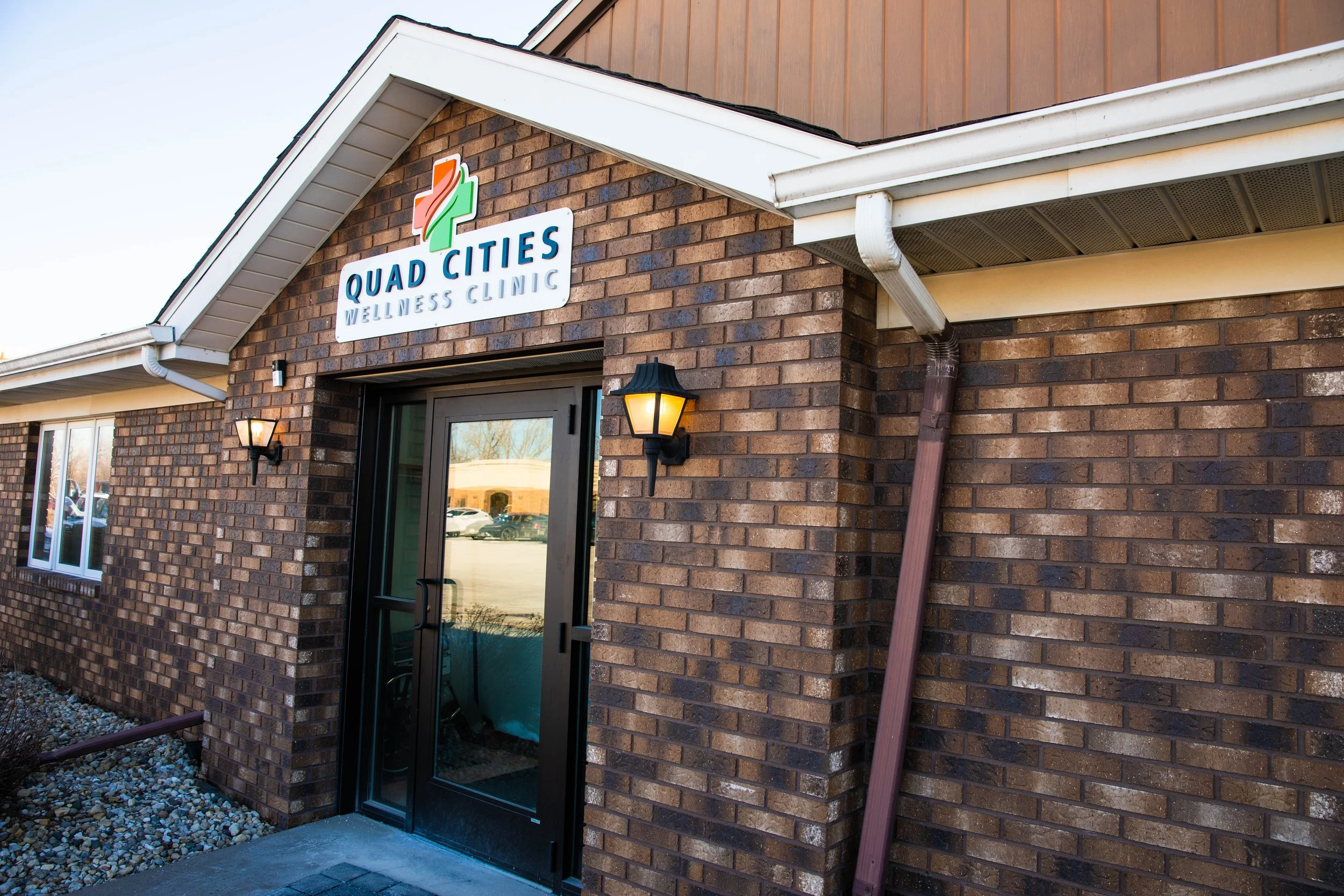 Exterior of Quad Cities Wellness Clinic, a brick building with a sign above the entrance, two wall-mounted lantern-style lights, glass door, and window, with gravel landscaping.