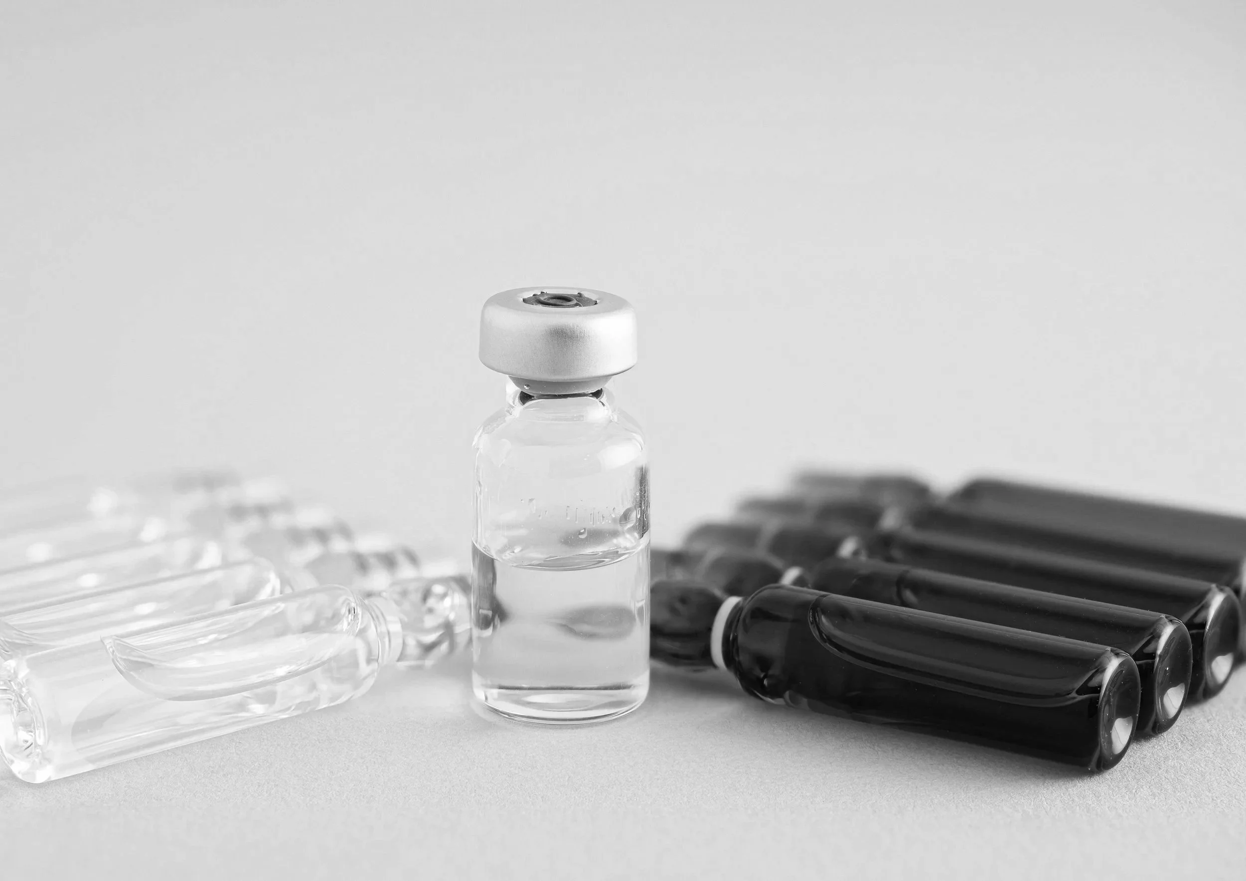 A small glass vial filled with a clear liquid, surrounded by several plastic ampoules containing a dark liquid, on a plain white background.
