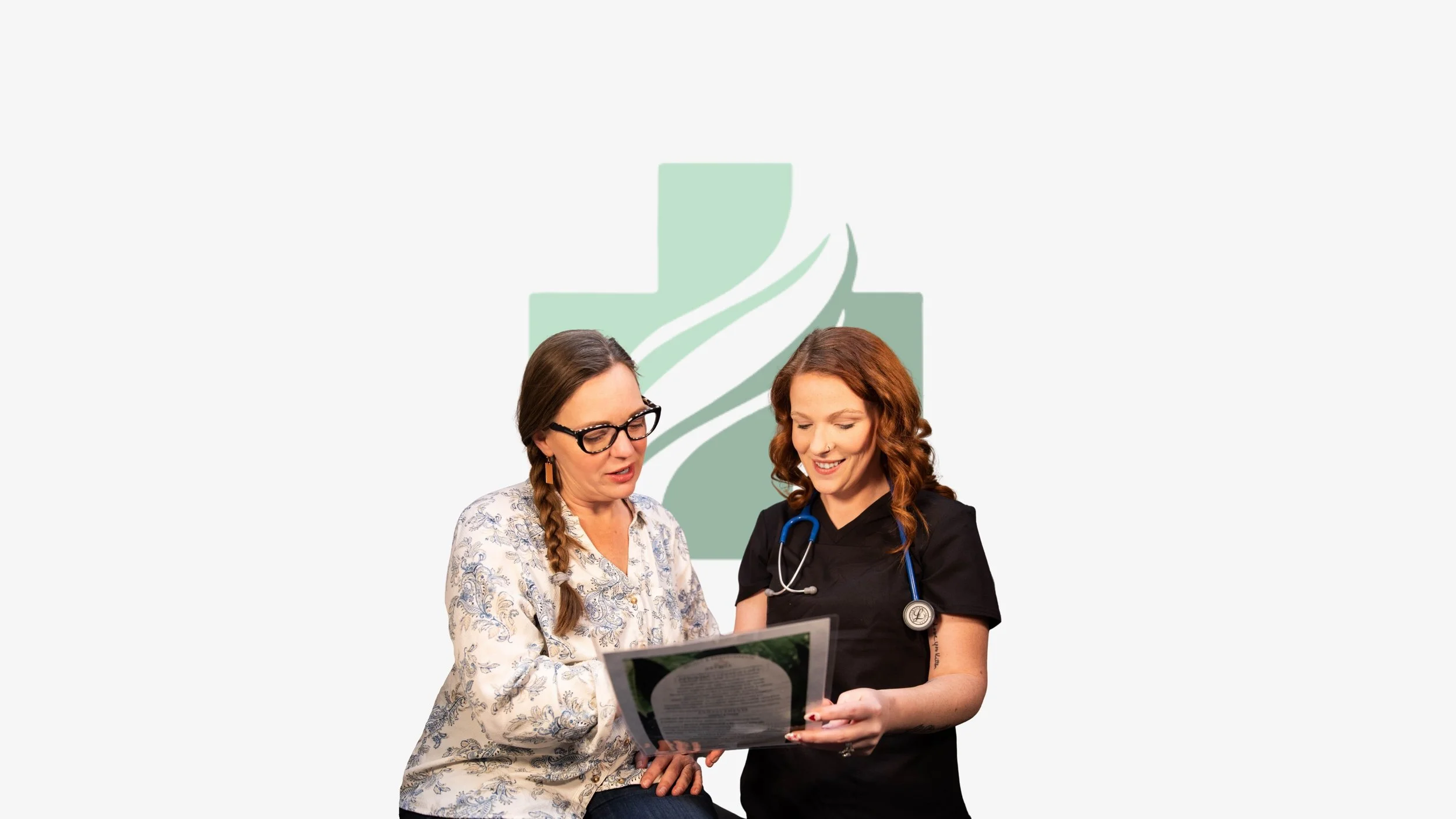 Two women, one with glasses and braided hair and the other with wavy red hair and a stethoscope, look at a document together with a medical cross and leaf logo in the background.