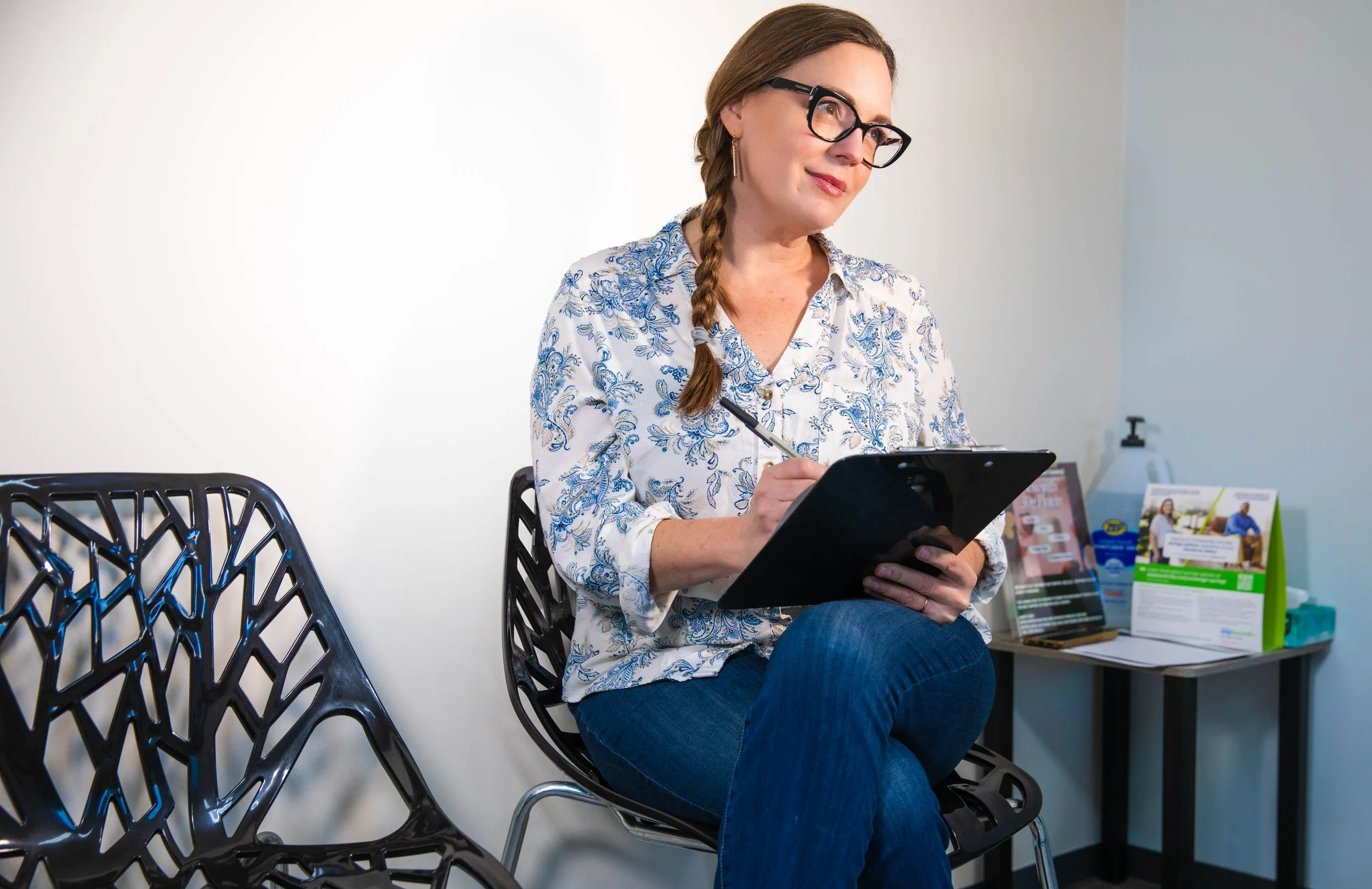 A woman with glasses and braided hair sitting in a waiting room, writing on a clipboard.