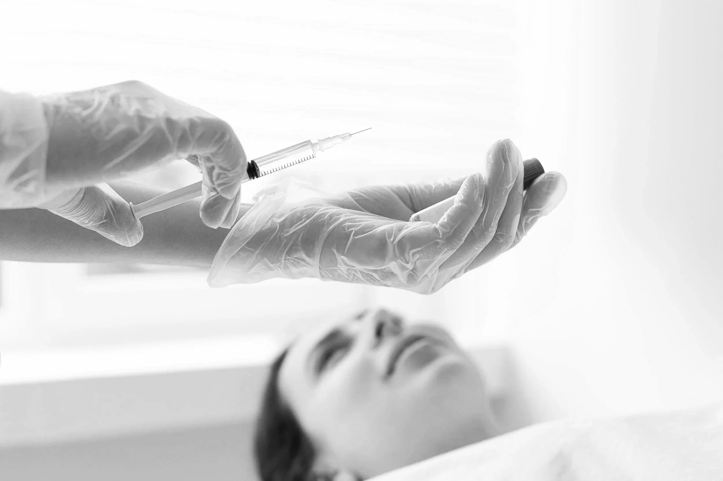 A healthcare worker wearing gloves and holding a syringe prepares an injection over a patient's arm in a clinical setting.