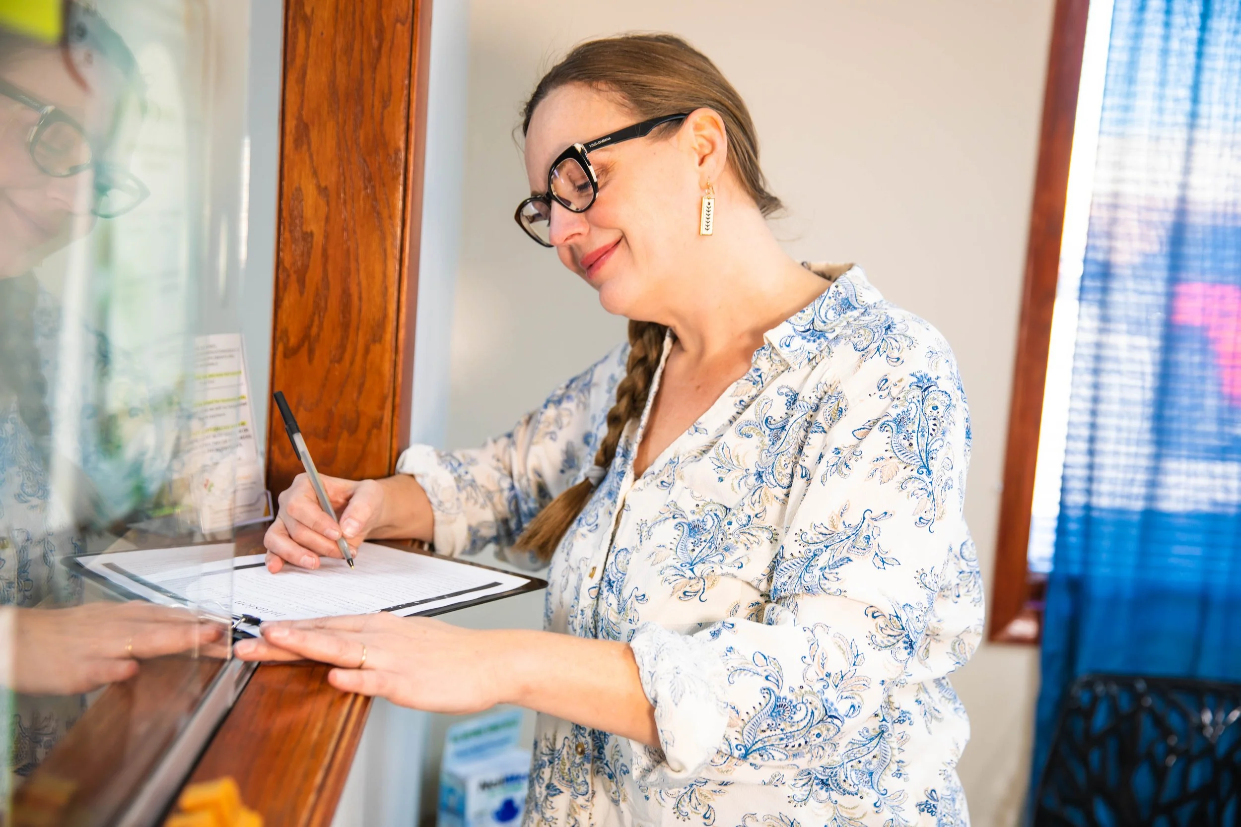 A woman wearing glasses and earrings, signing a document at a reception desk with a pen.