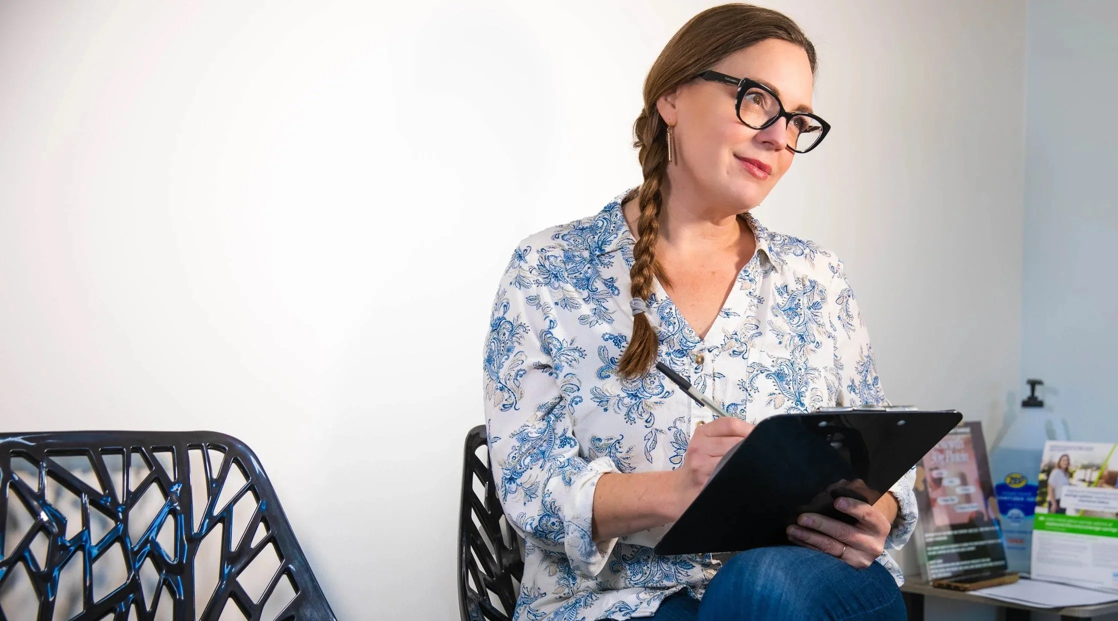 A woman with brown hair in a braid, wearing glasses and a white blouse with blue floral patterns, sitting and taking notes on a clipboard with a pen. She appears to be in a professional office setting.
