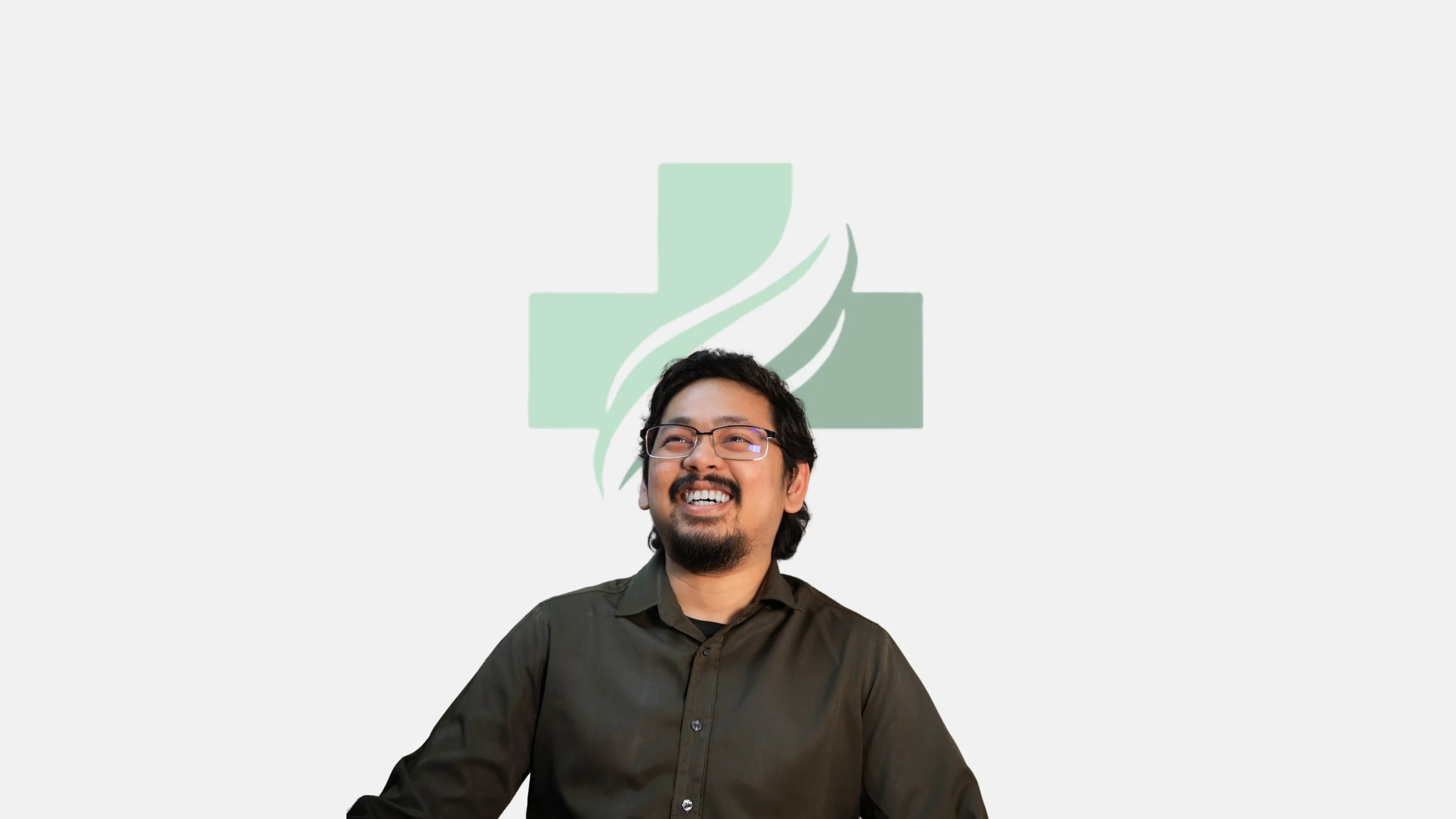 A man with glasses smiling, wearing a dark shirt, sitting in front of a light gray background with a green medical cross logo and a white flame design.