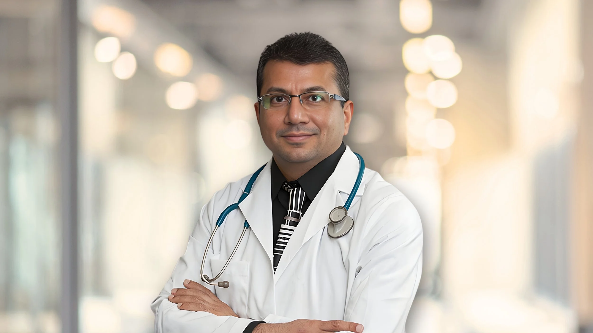 A male doctor wearing glasses, a white lab coat, and a stethoscope around his neck, standing with arms crossed in a brightly lit medical facility hallway.