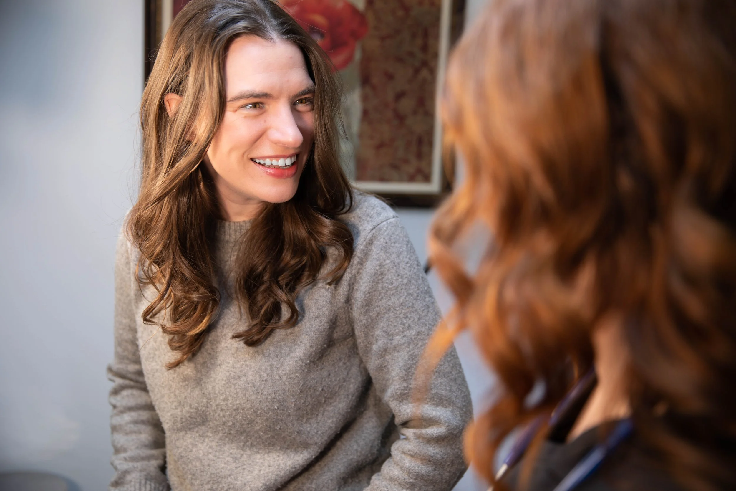 Two women engaged in a friendly conversation, smiling at each other indoors.