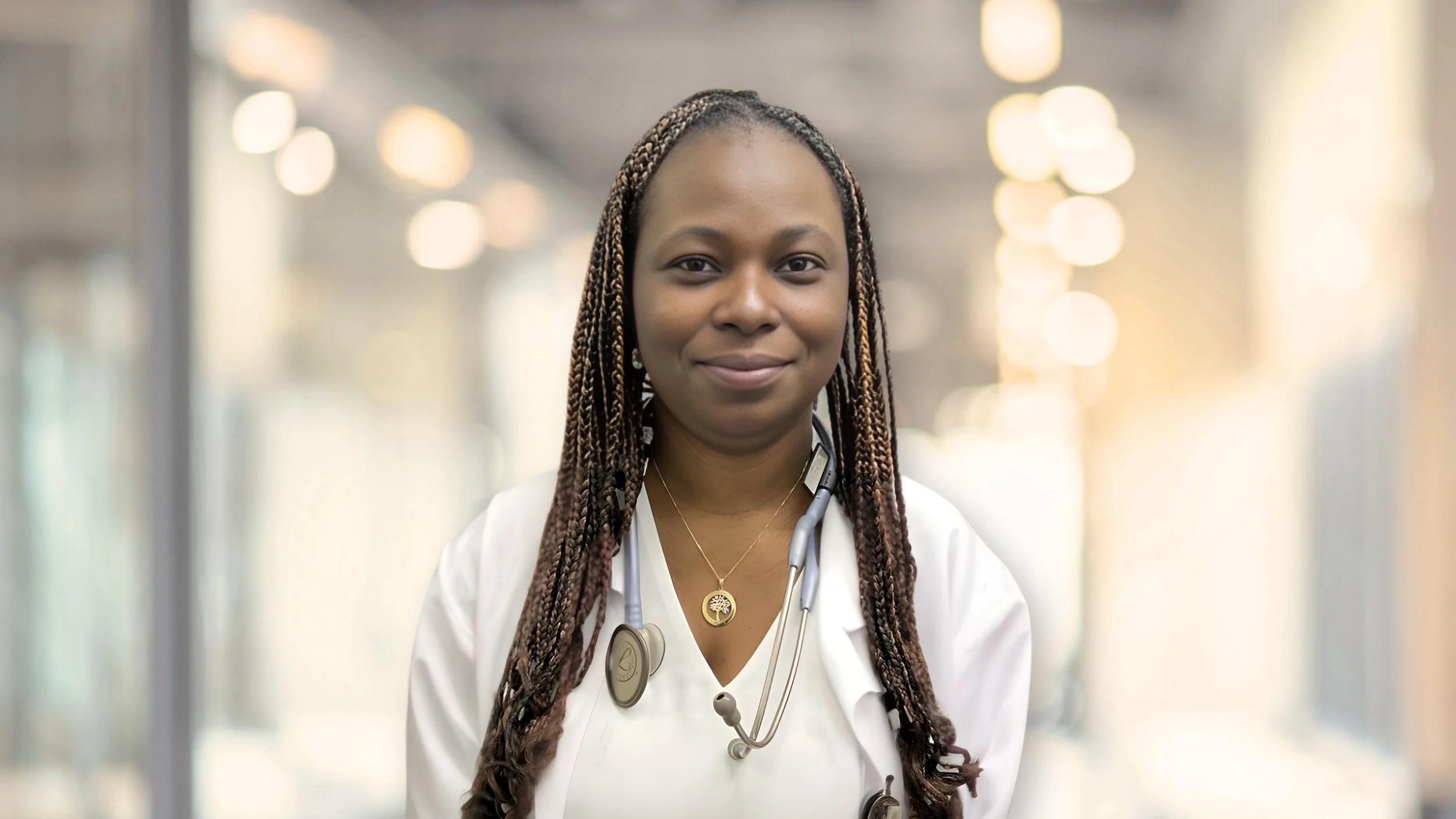 A female healthcare professional with dreadlocks wearing a white lab coat and stethoscope, standing in a bright, modern medical facility or hospital corridor.