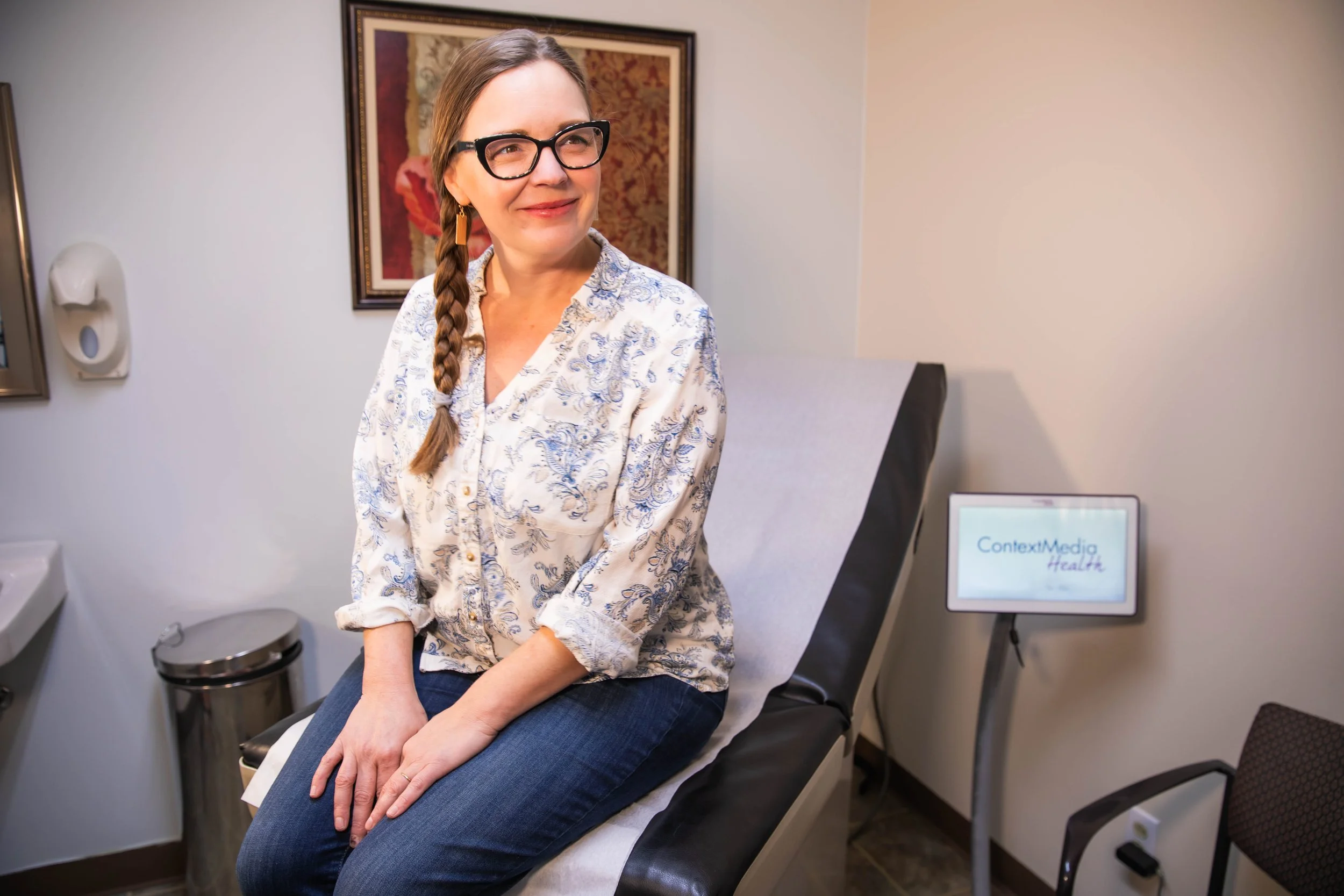 A woman sitting on an examination table in a medical office, smiling and looking to the side, wearing glasses and a floral blouse.
