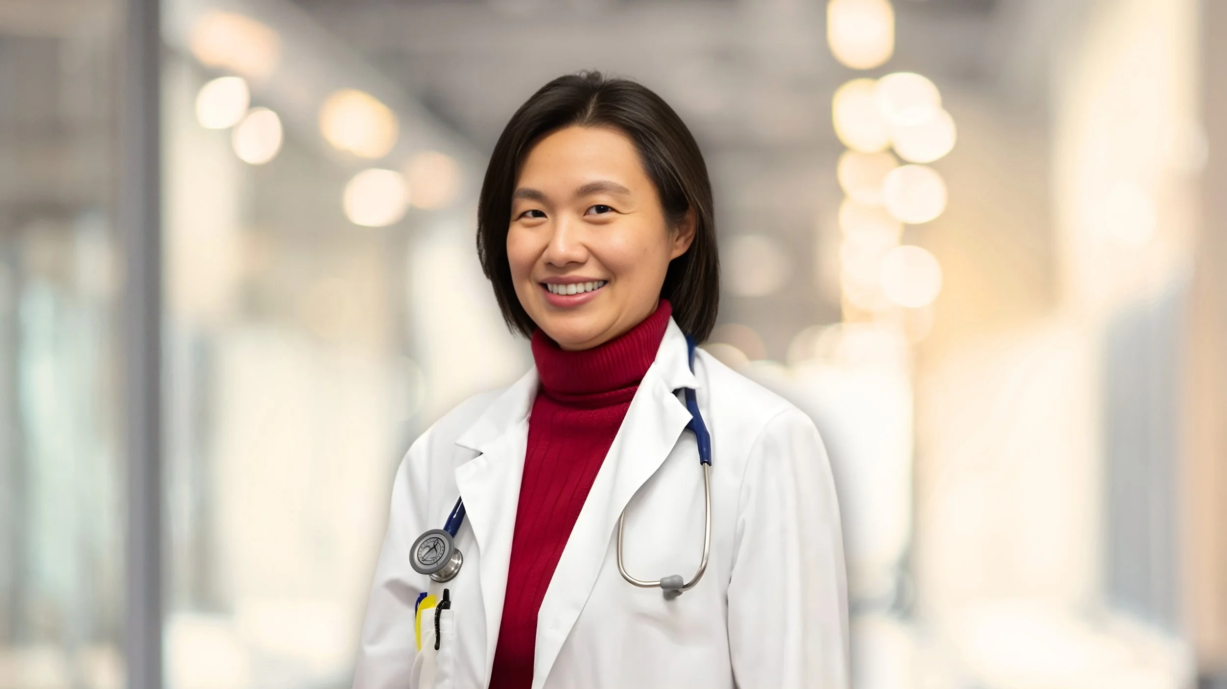 A female doctor with short black hair wearing a white lab coat, a red turtleneck sweater, and a stethoscope around her neck, smiling in a well-lit hospital corridor.