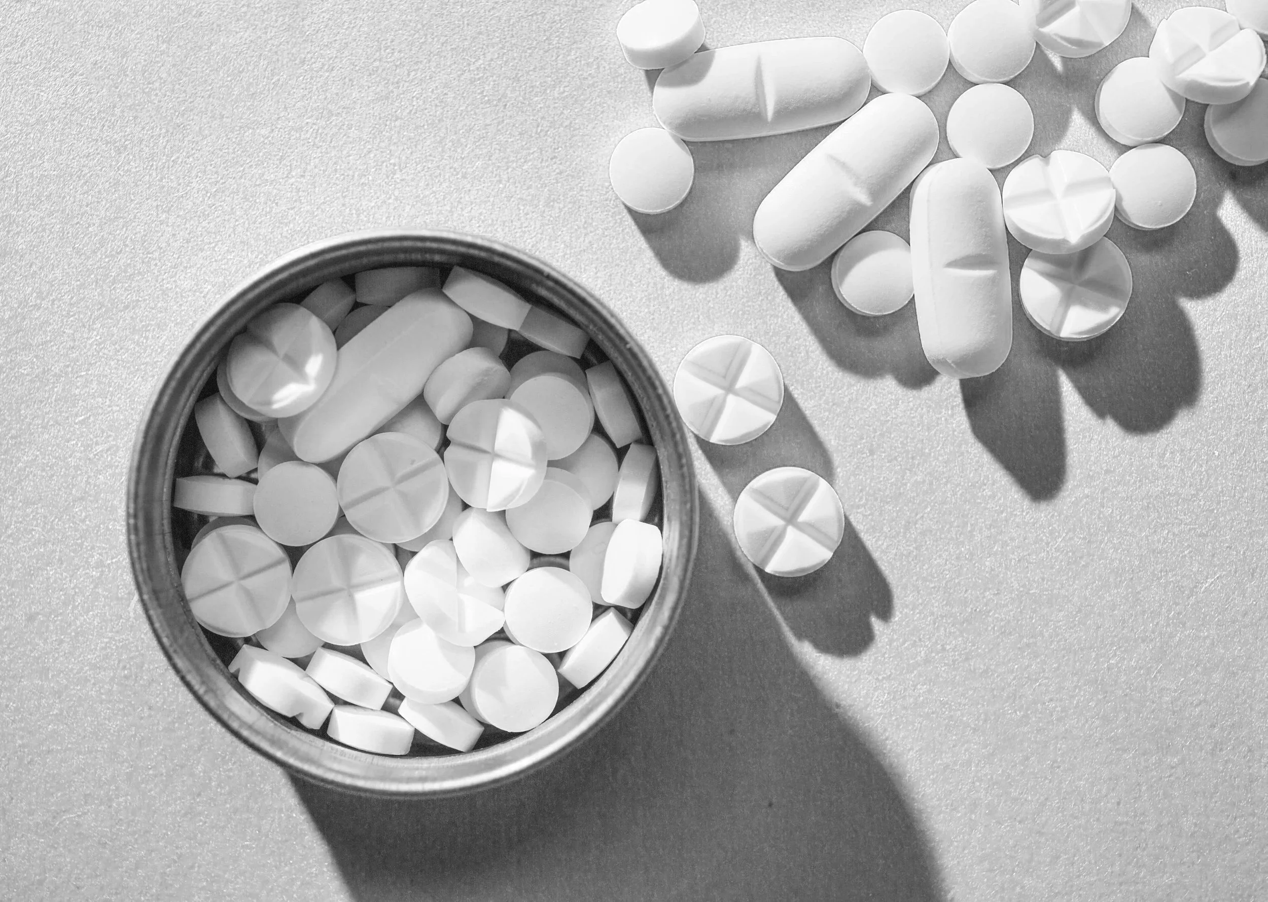 A round open container filled with various white pills and tablets on a flat surface, with some pills spilled outside the container.