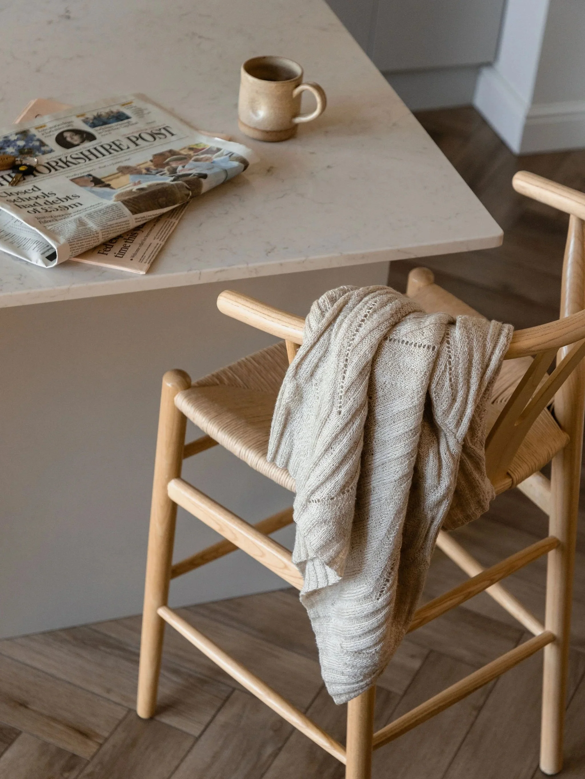 A wooden chair with a beige knitted blanket draped over the backrest, positioned next to a kitchen counter with a coffee mug, a folded newspaper, and keys on its surface, and a wooden floor below.