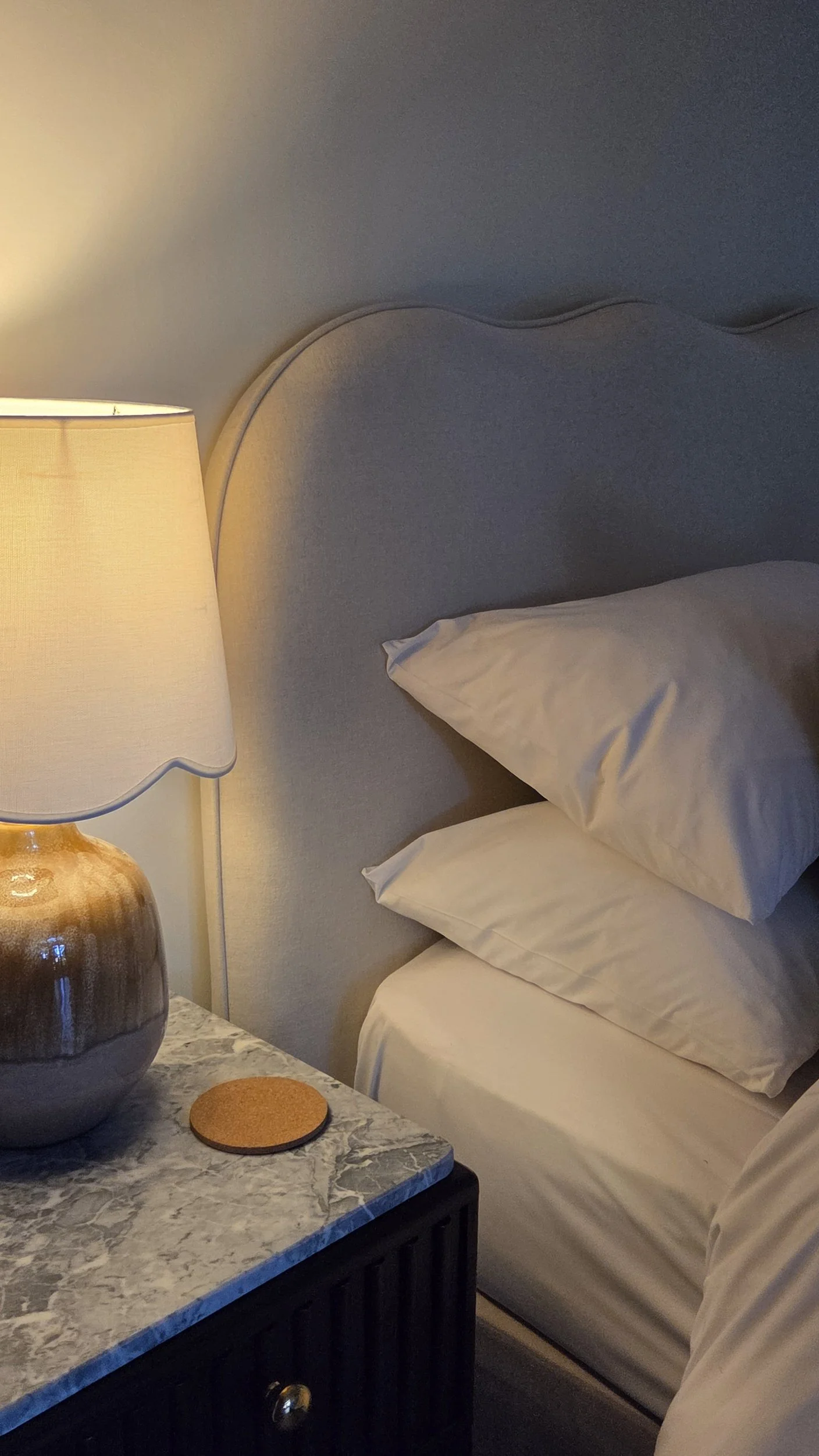 A bedroom scene with a bedside table holding an open book and a lamp, along with a bed with pillows and a quilt, illuminated by warm lighting.