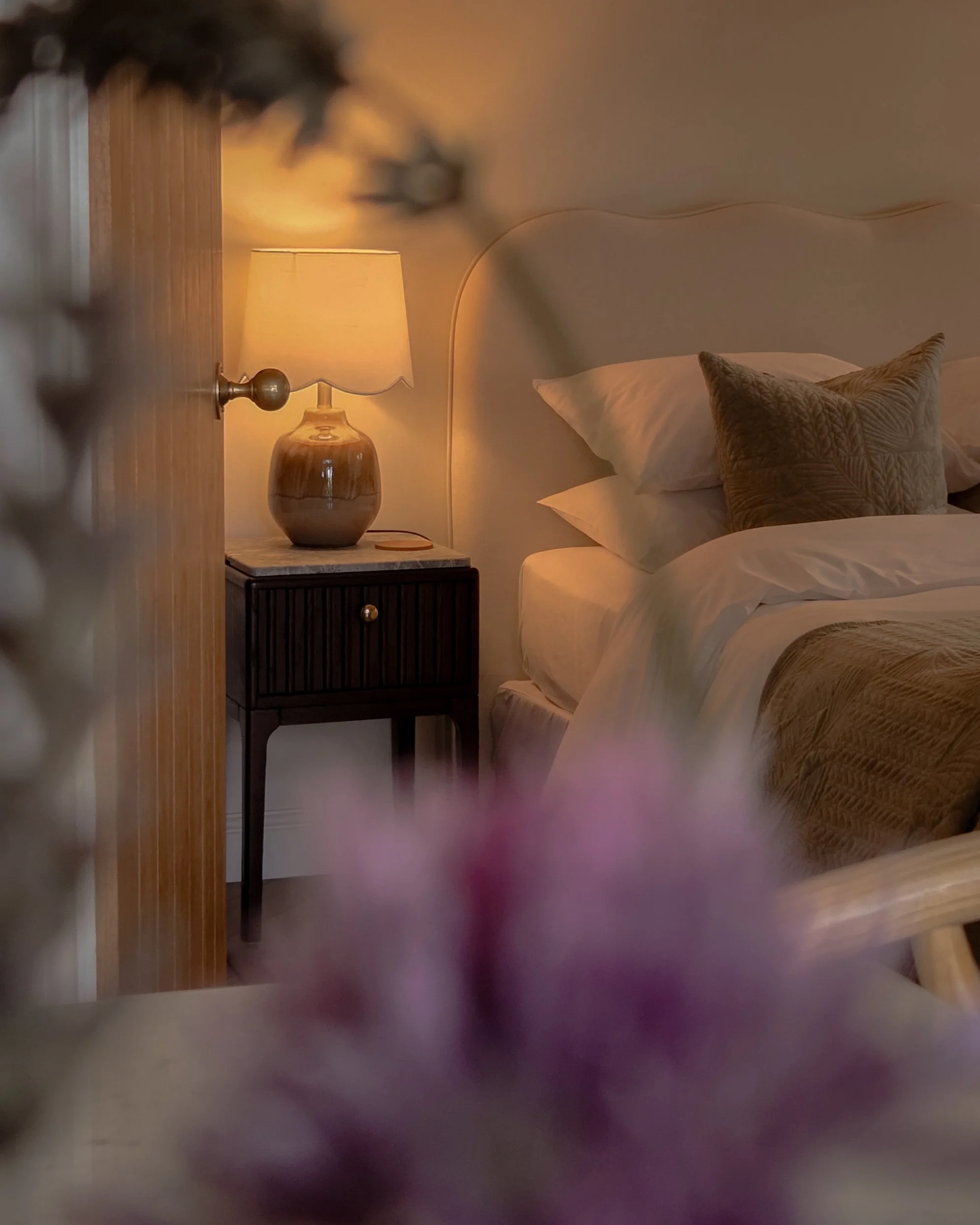 A wooden door, a bedside table with a marble top and a ceramic lamp, a bed with white sheets, pillows, and a textured throw pillow, and a wall with a curved line and a round mirror or clock.