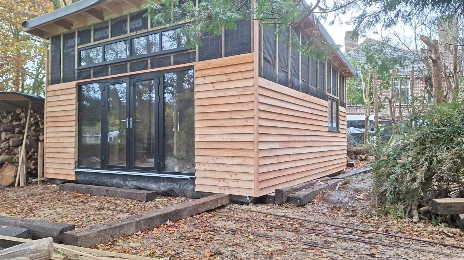 A house under construction with wooden siding on the lower level and black-framed glass doors and windows, surrounded by trees and construction materials.