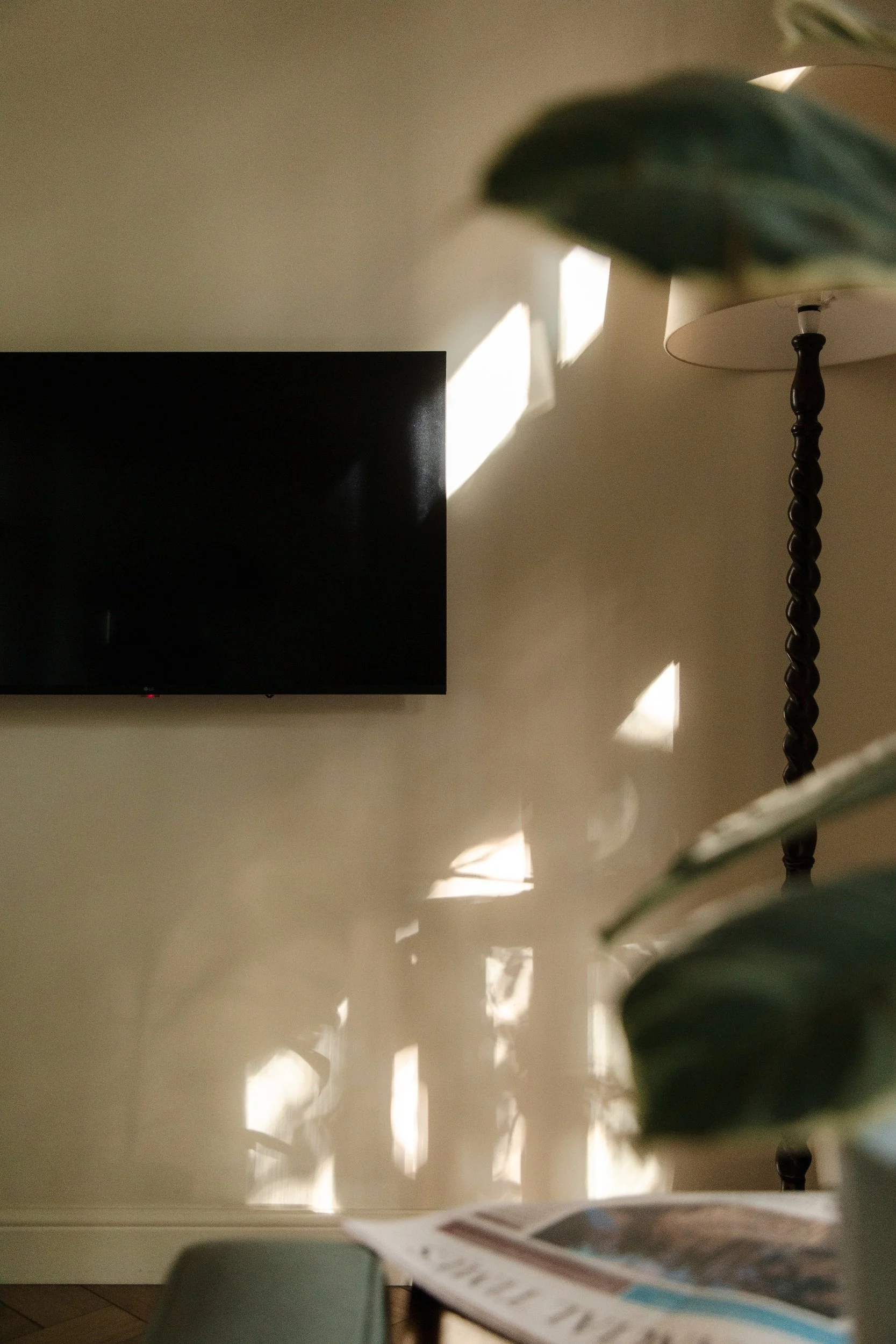Sunlight creating rectangular shadow patterns on a cream-colored wall with a flat-screen TV mounted on it, and a tall lamp with a twisted dark wood stand visible on the right. Part of a blurred newspaper or magazine is at the bottom of the image.