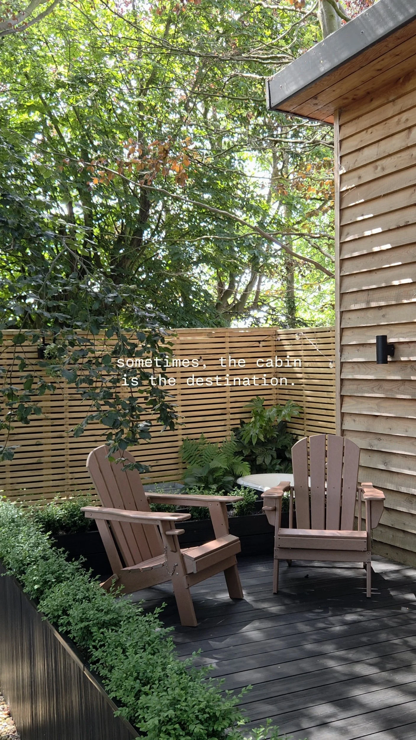 A cozy outdoor patio with wooden Adirondack chairs, lush green plants, and a bamboo privacy fence, surrounded by tall trees. The scene is peaceful and inviting, with sunlight filtering through the leaves.