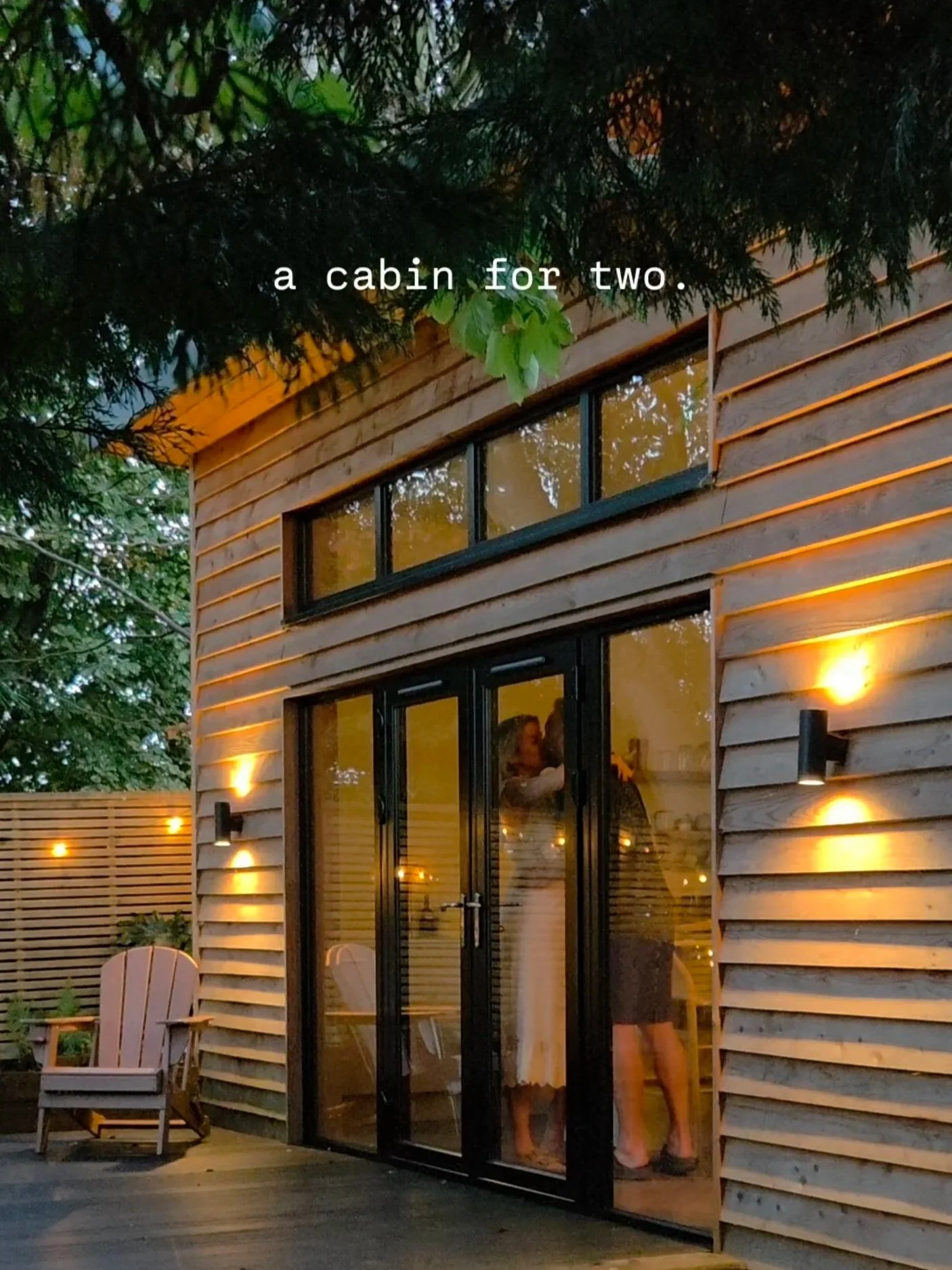 A cozy wooden cabin with large glass doors and warm exterior lighting, surrounded by green trees and outdoor chairs.