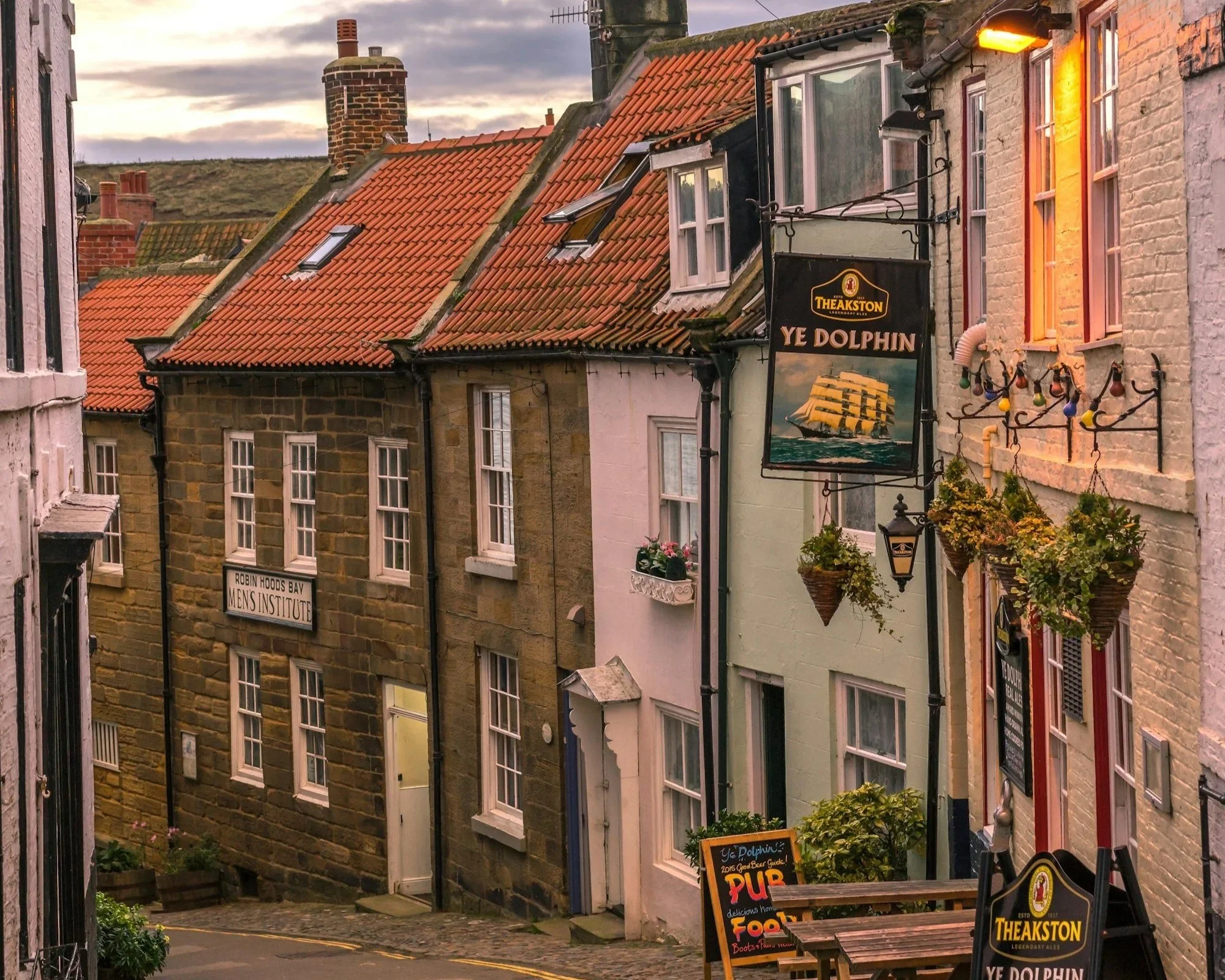 Picturesque street view showing old brick and stone buildings with windows, signs for Ye Dolphin pub, hanging light fixtures, flower baskets, and a somewhat cloudy sky at sunset.