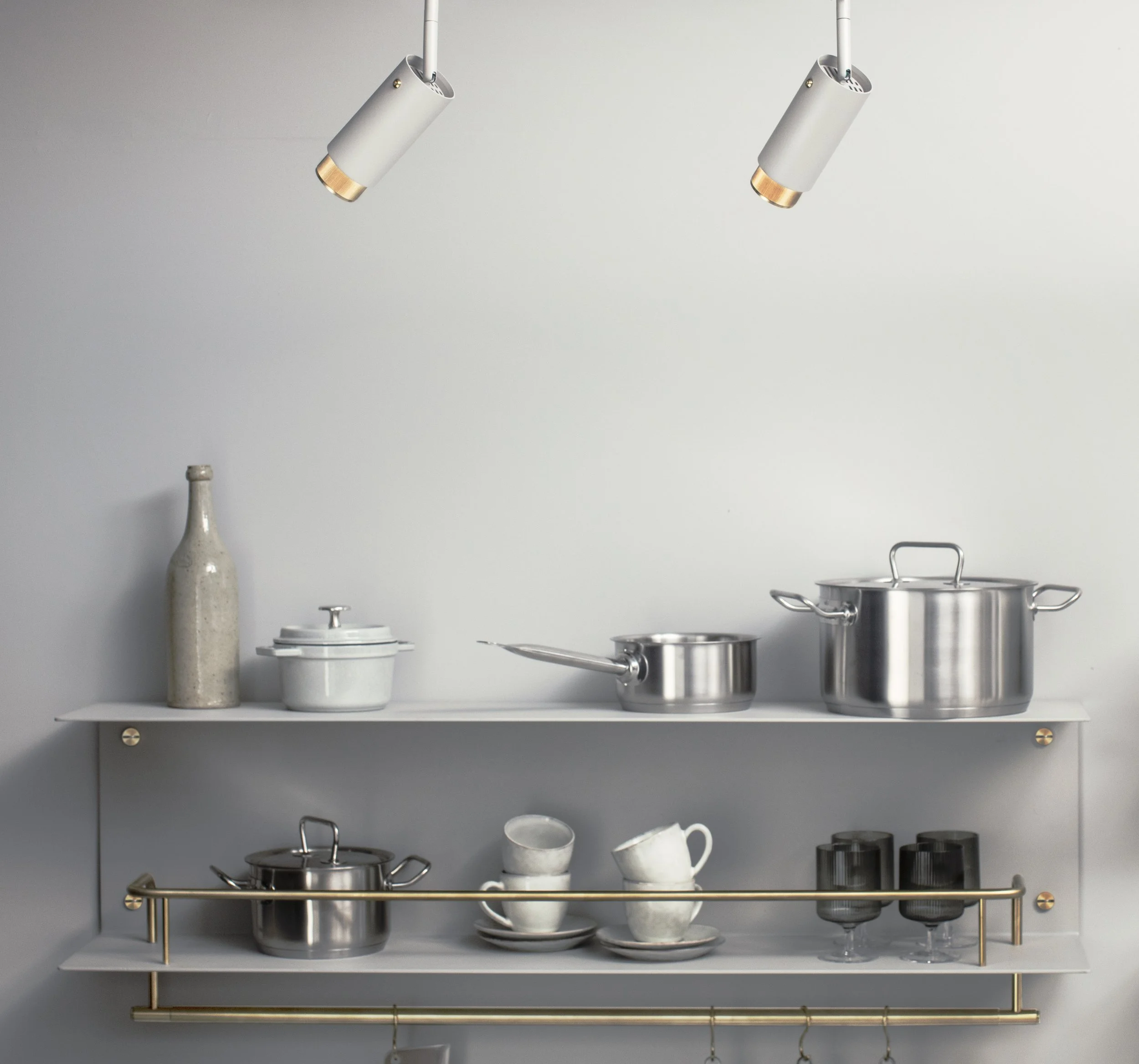 Kitchen shelf with white and stainless steel pots, dishes, mugs, and a bottle on a minimalist rack against a light gray wall.