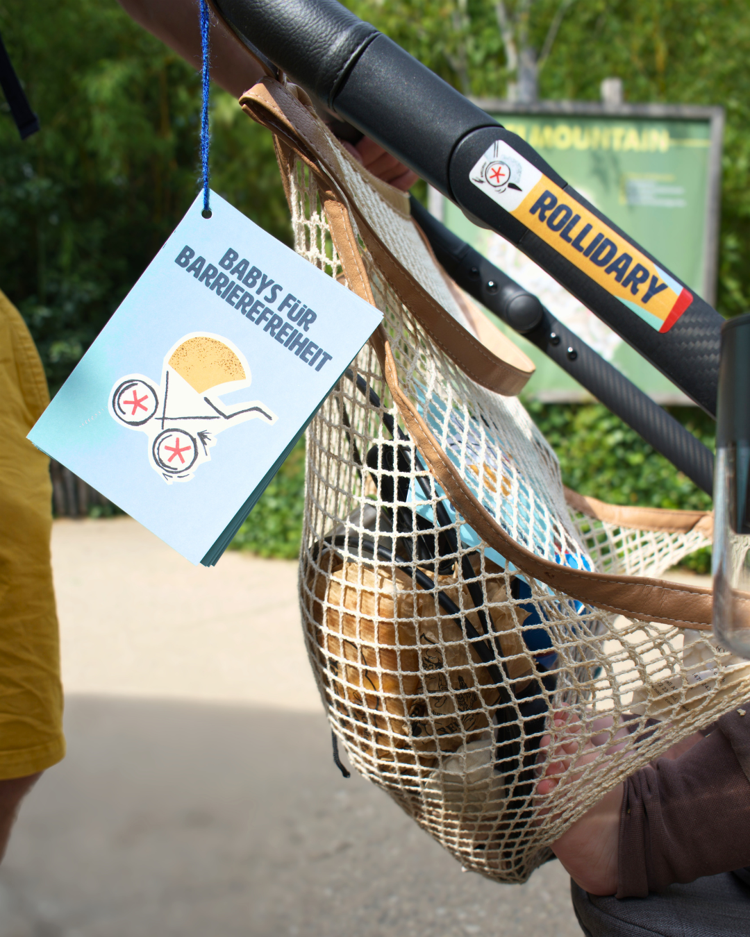 An einem Kinderwagen hängen Postkarten von „Babys für Barrierefreiheit“; am Griff klebt ein #Rollidary-Aufkleber.