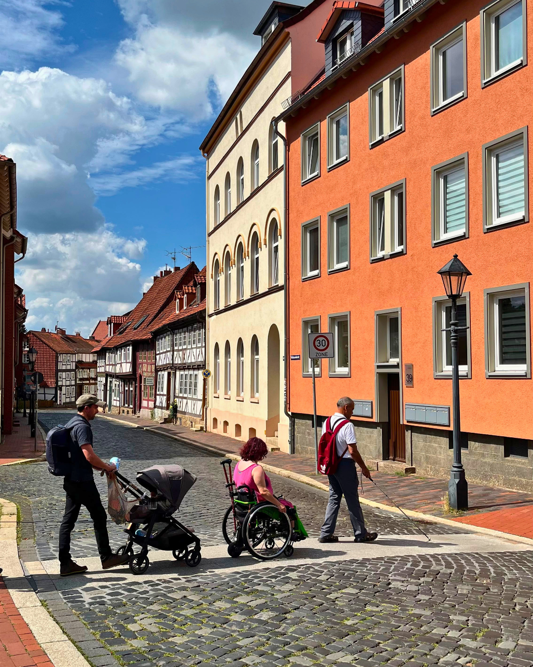 Ein Mann mit Kinderwagen, eine Frau im Rollstuhl und ein Mann mit Langstock überqueren eine Kopfsteinpflasterstraße in Hildesheim.