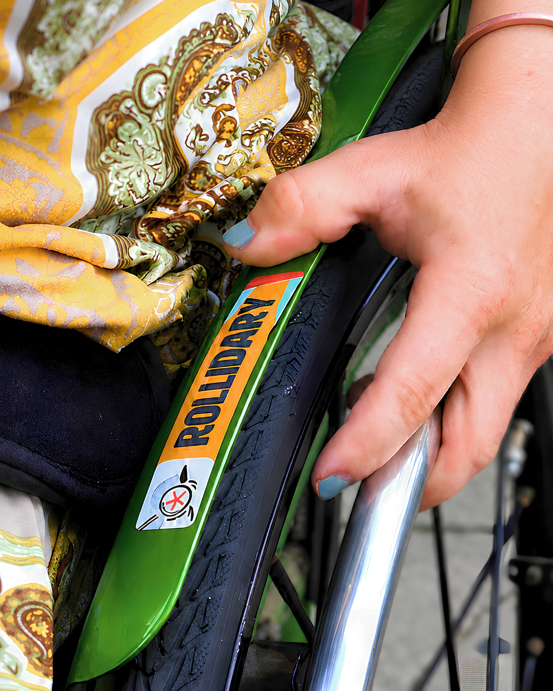Nahaufnahme einer Hand am Greifreifen eines Rollstuhls; am grünen Rahmen klebt ein gelber #Rollidary-Aufkleber.