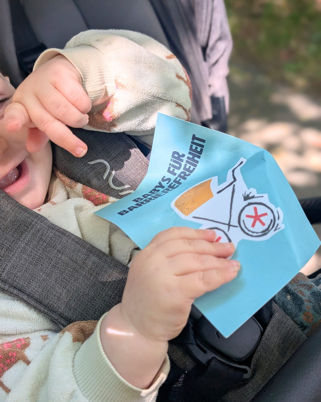 Ein Baby im Kinderwagen hält eine blaue Postkarte mit der Aufschrift „Babys für Barrierefreiheit“.
