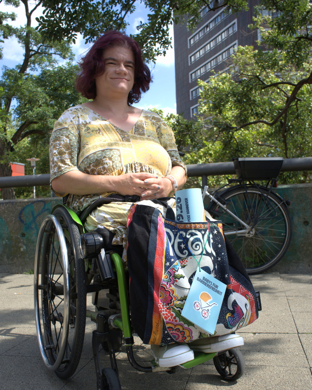Mona Bernhard sitzt im Rollstuhl; an ihrer Tasche hängt eine Postkarte von „Babys für Barrierefreiheit“.