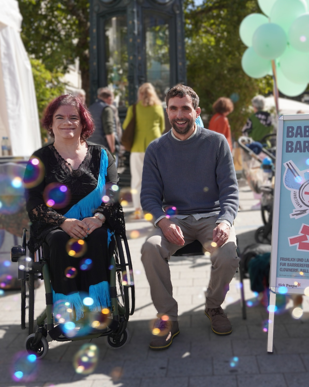 Die Organisator*innen der Demo, Mona Bernhard und Max Balzer, auf der Demo "Babys für Barrierefreiheit" am Entdeckerfest. Im Vordergrund schillern Seifenblasen.