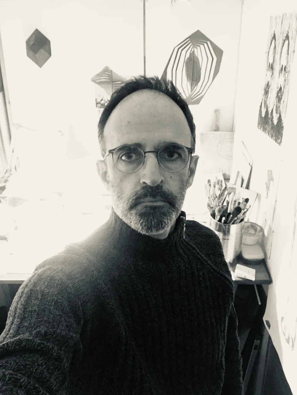 A man with glasses and a beard taking a selfie in an art studio, surrounded by various artworks and art supplies.