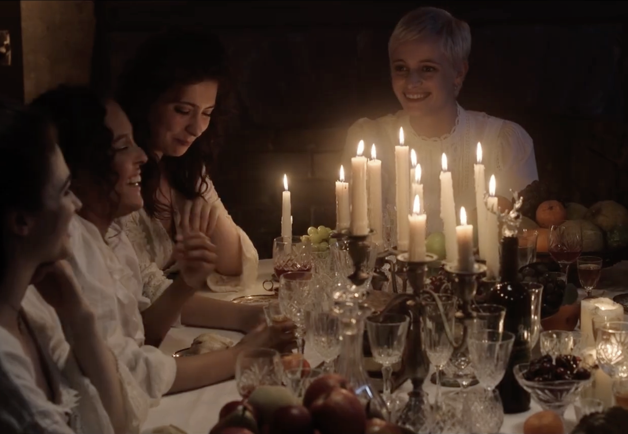 A group of women gathered around a dinner table with lit candles, enjoying a celebration in warm lighting, with various fruits and glassware on the table.