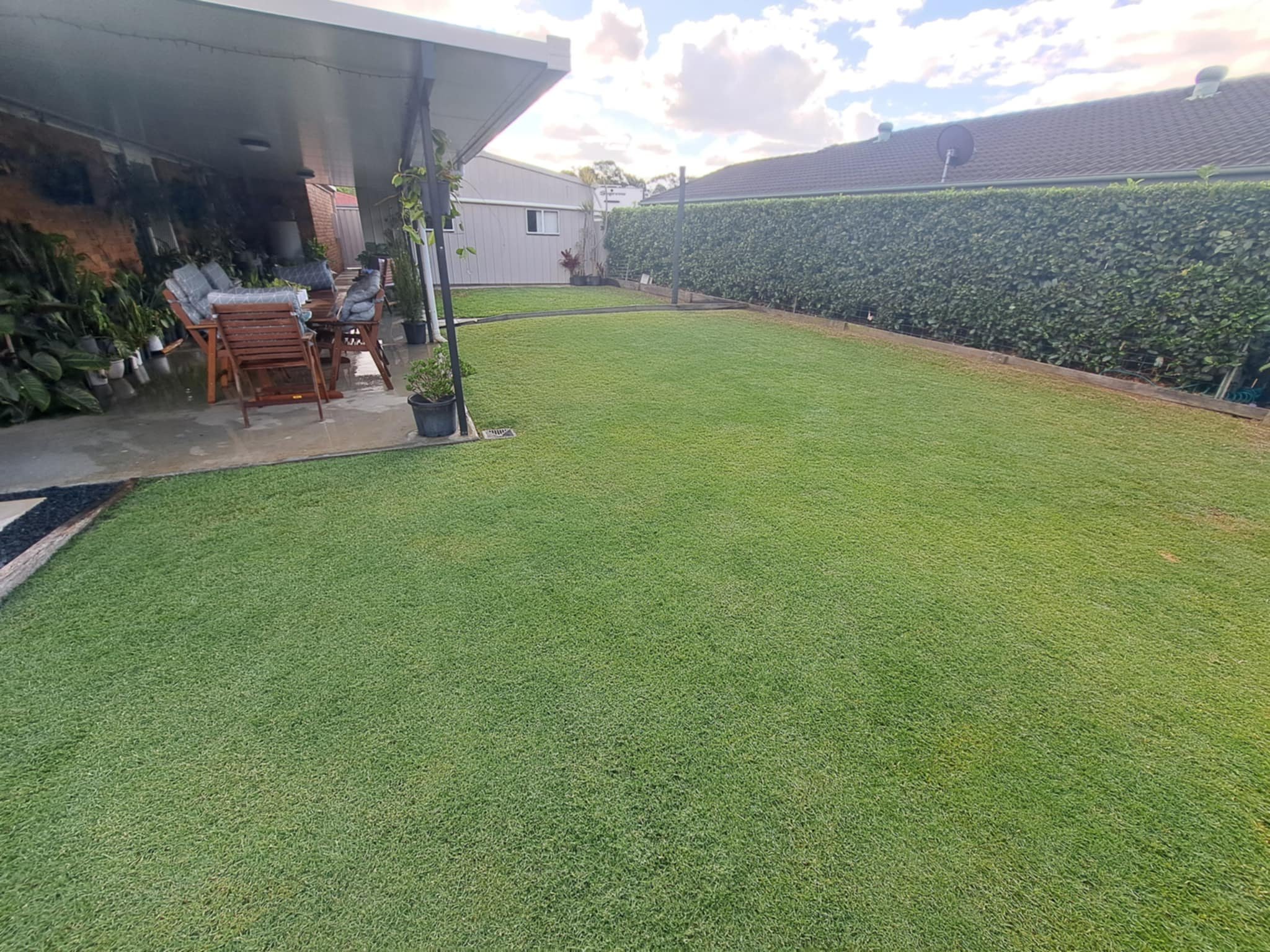 Backyard with a well-maintained lawn, patio with outdoor furniture, potted plants, a hedge on the right, and a house with satellite dishes on the roof in the background.