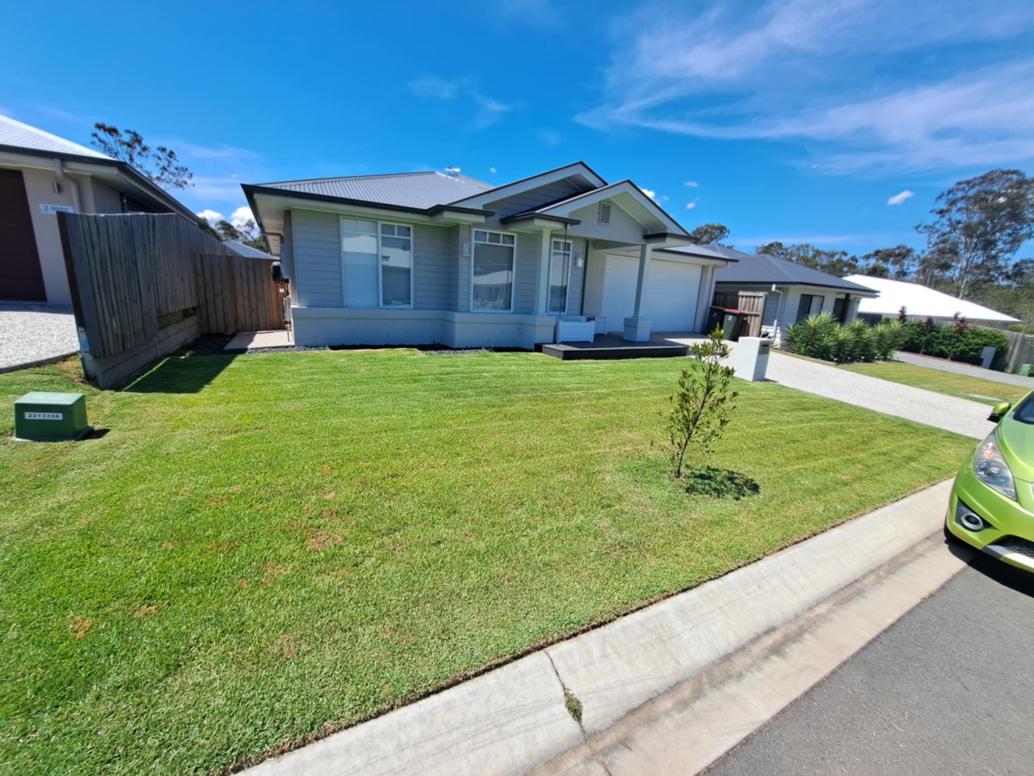 Front yard of a modern house with a well-maintained lawn, small young tree, concrete pathway, parked green car, and neighboring houses under a blue sky with some clouds.