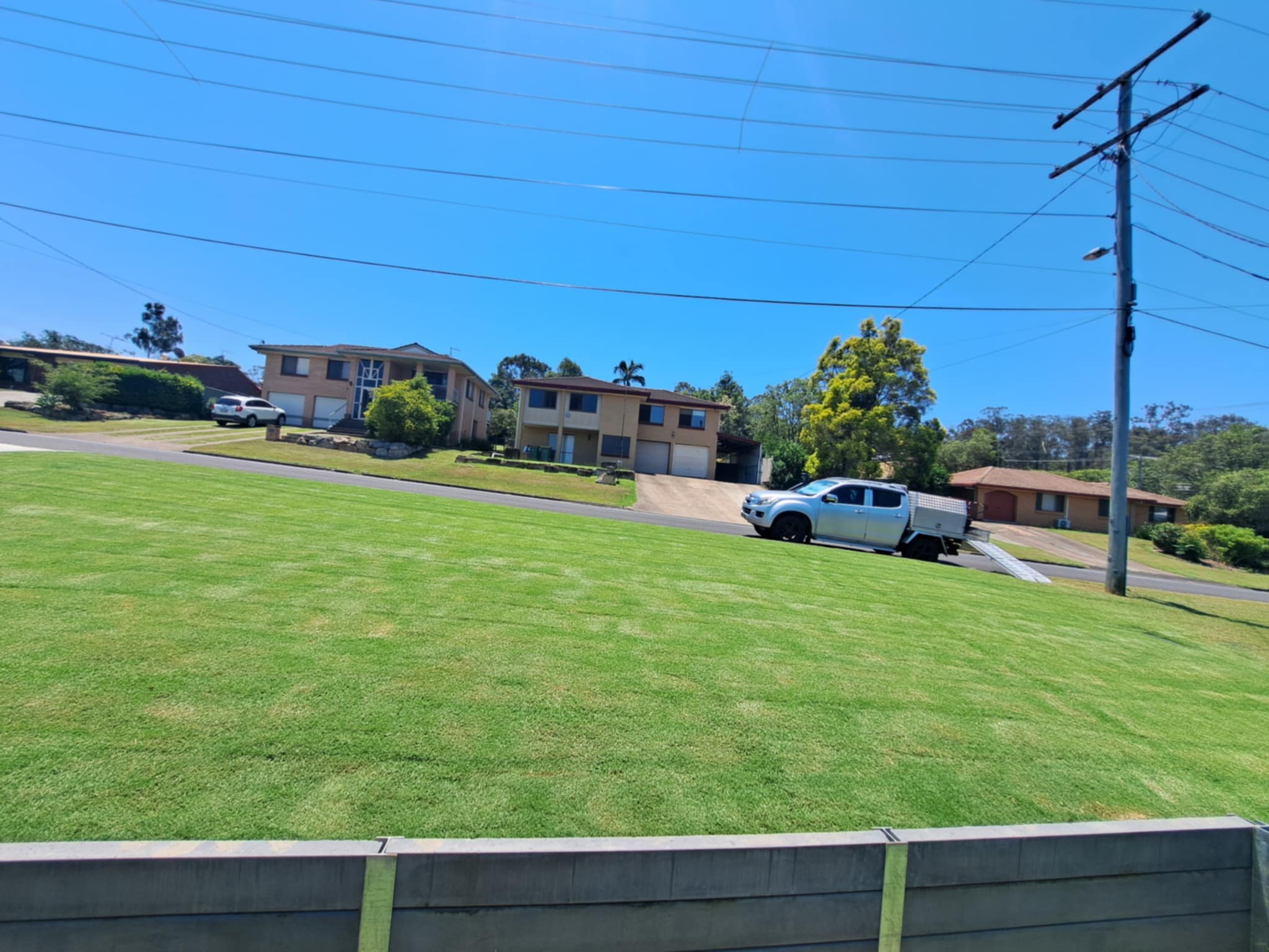 A grassy hill with a street and houses in the background, a white pickup truck parked on a ramp, and power lines overhead under a clear blue sky.