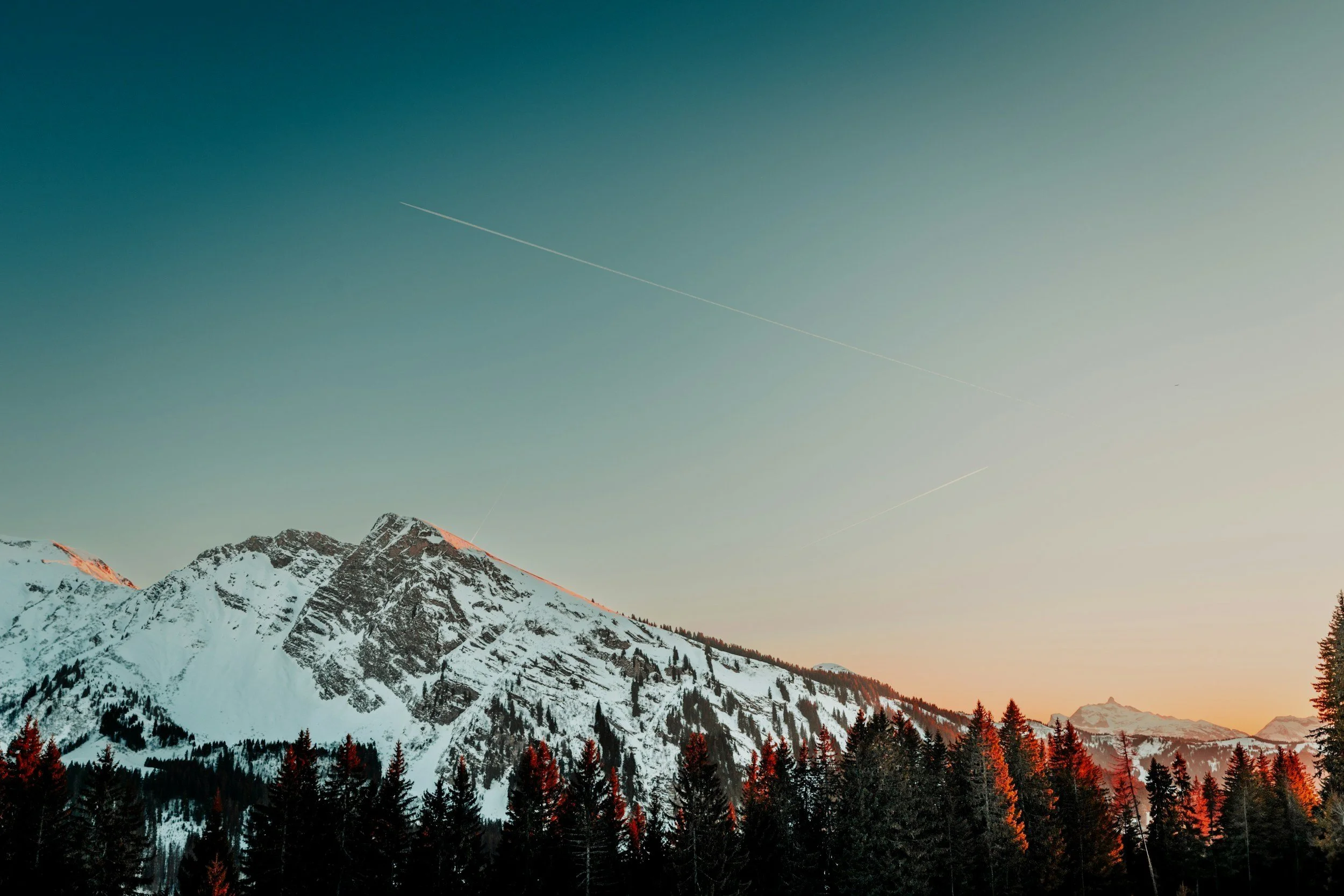 Snow-covered mountain range at sunset with forest in the foreground and contrails in the sky.