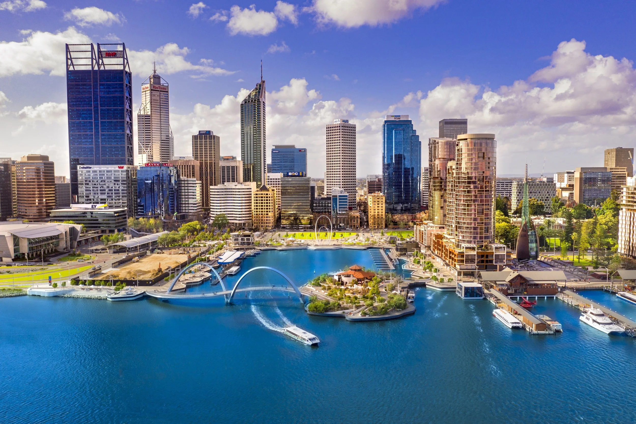Skyline of downtown Perth with high-rise buildings, a river with boats, and a park area in the foreground.