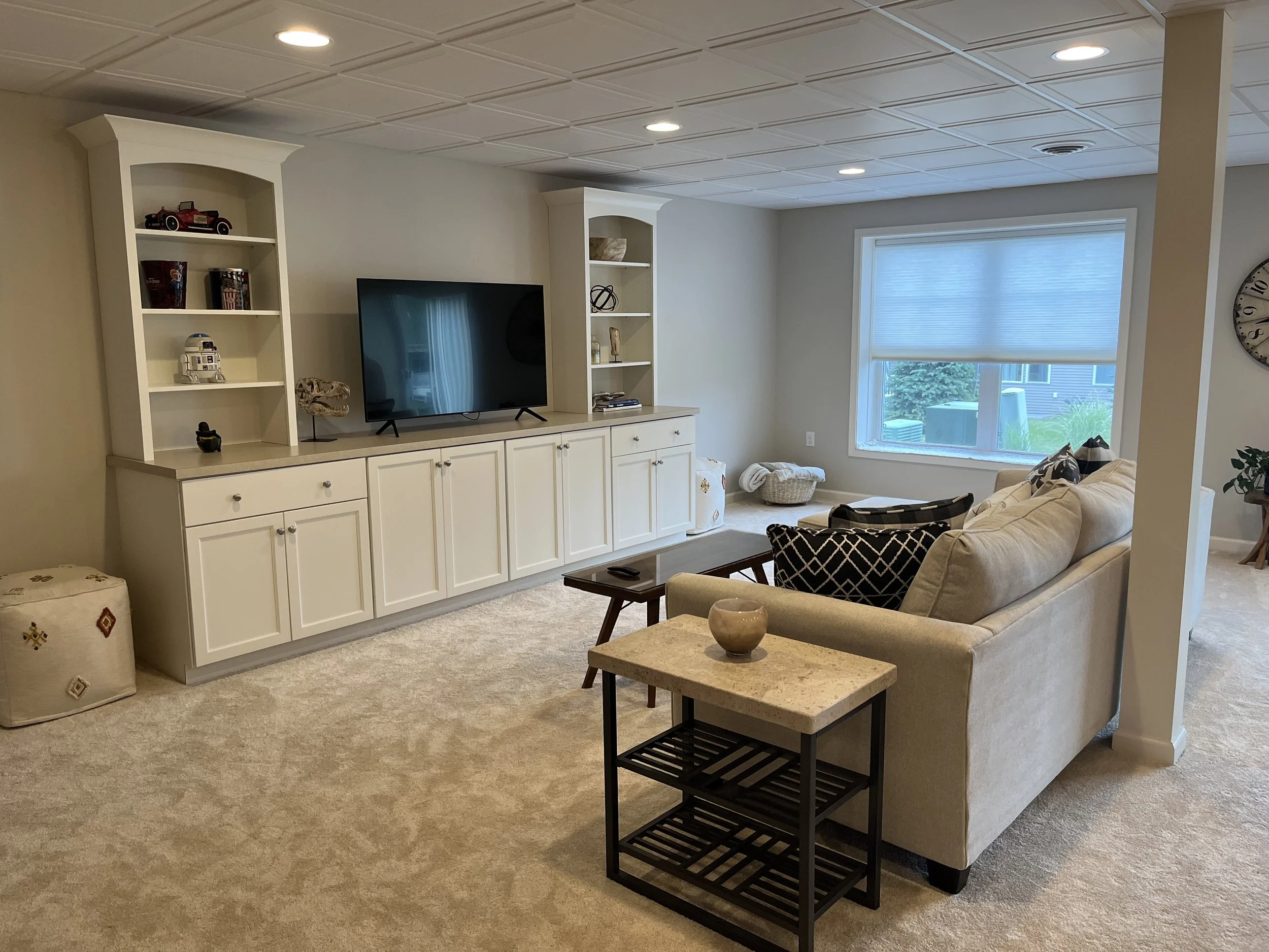 A living room with a beige carpet, cream-colored sofas, a white cabinet with shelves and a TV, a small coffee table with a decorative bowl, a window with blinds, and various decorative items on the shelves.