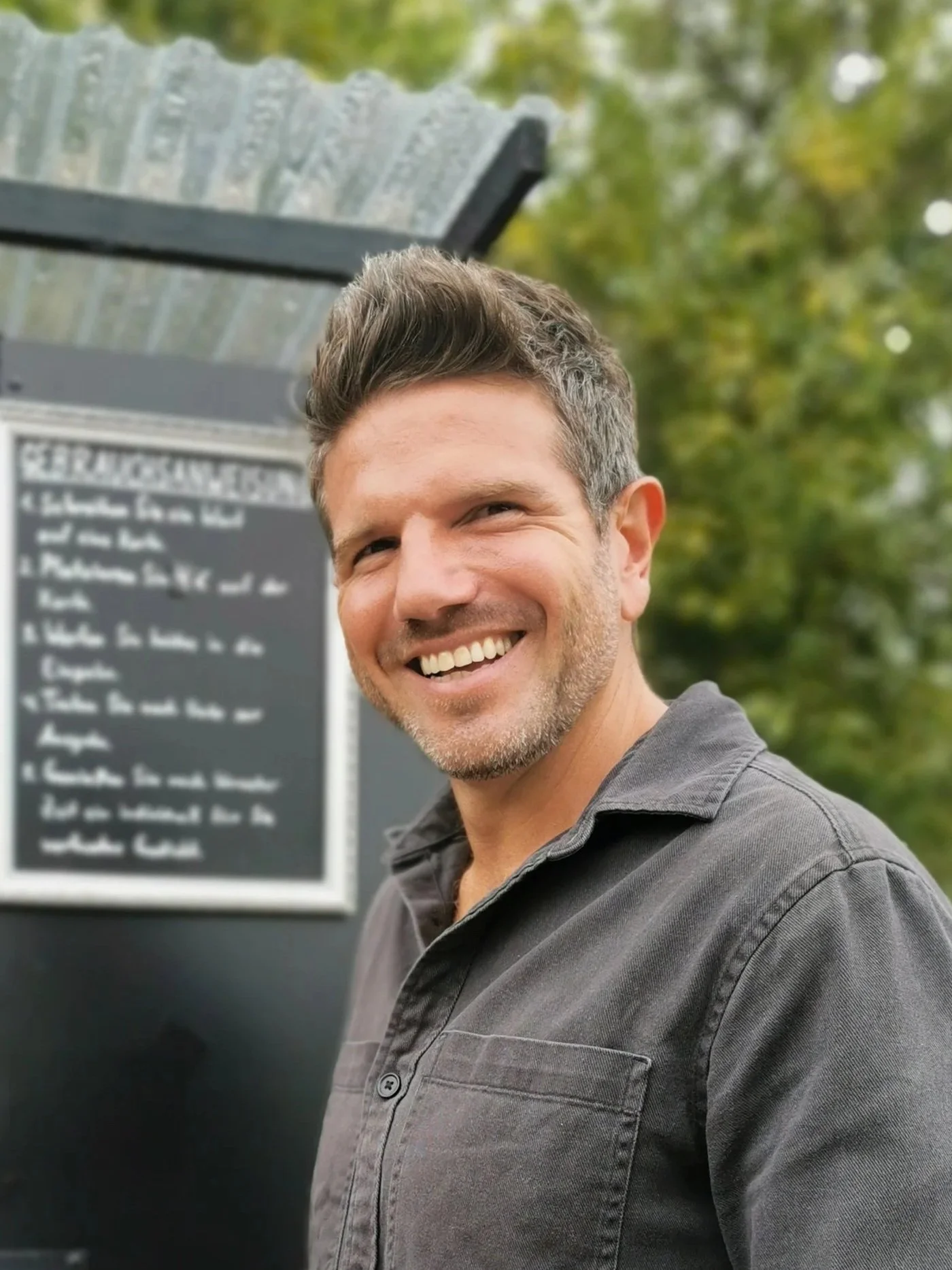 Ein lachender Mann mit dunklem Shirt vor einer Tafel im Freien, mit Bäumen im Hintergrund.