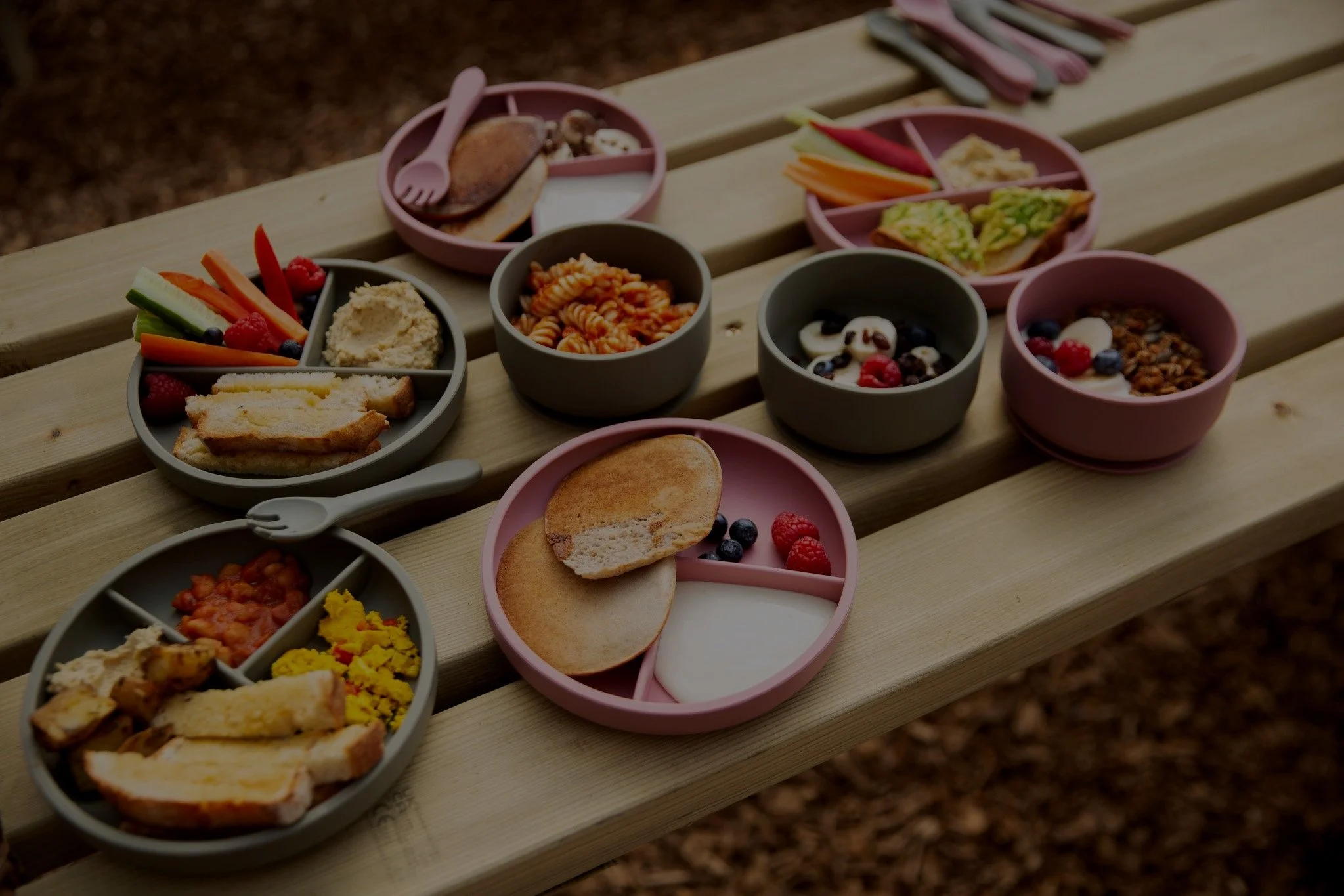 Picnic table with assorted children's meal plates, featuring pancakes, fruits, toast, hummus, vegetables, pasta, baked beans, and yogurt with berries and granola. Utensils are placed nearby.