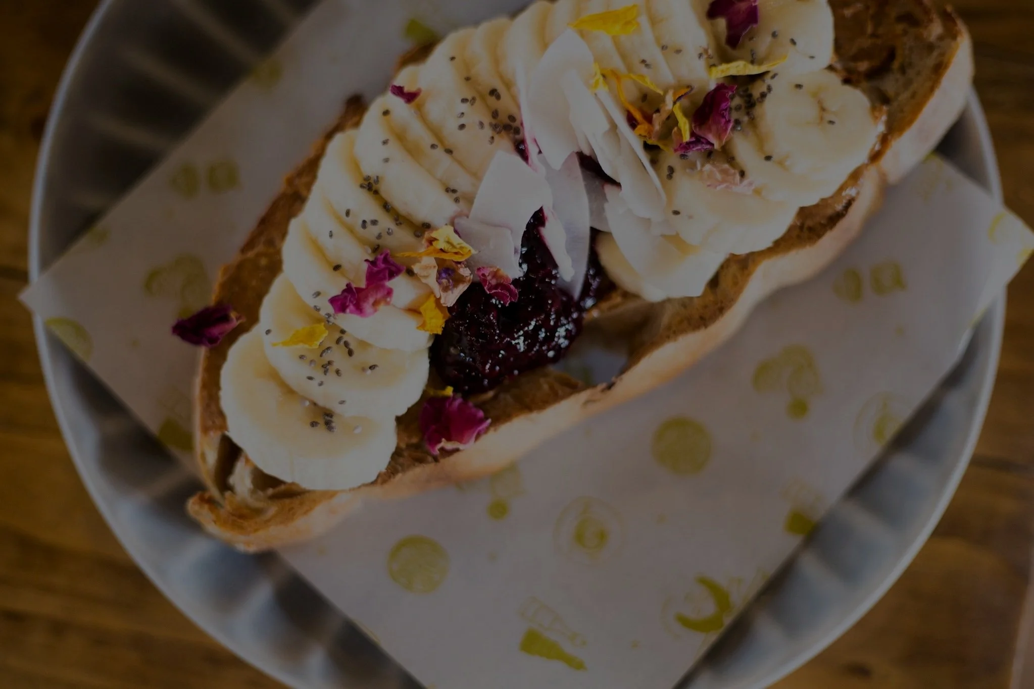 Open-faced sandwich with banana slices, chia seeds, jelly, and flower petals on a plate.