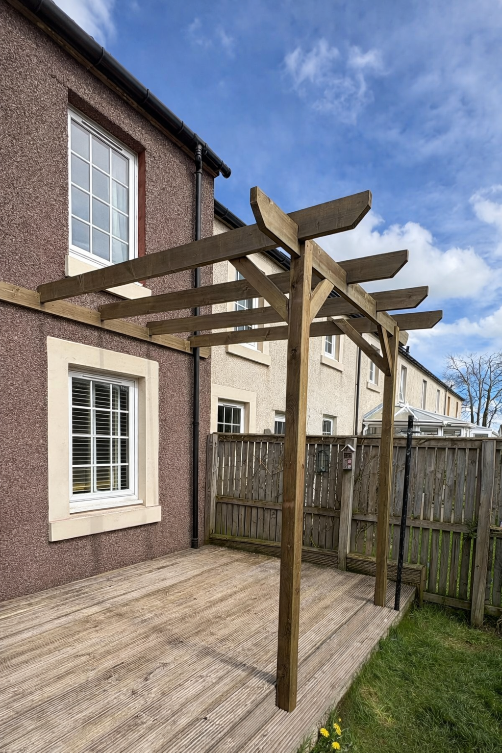 A new wooden pergola frame is being built on a backyard deck next to a house with textured brown and beige exterior walls and white-framed windows. The wooden structure is set on a weathered wooden deck and surrounded by a fenced backyard with some grass and yellow flowers.