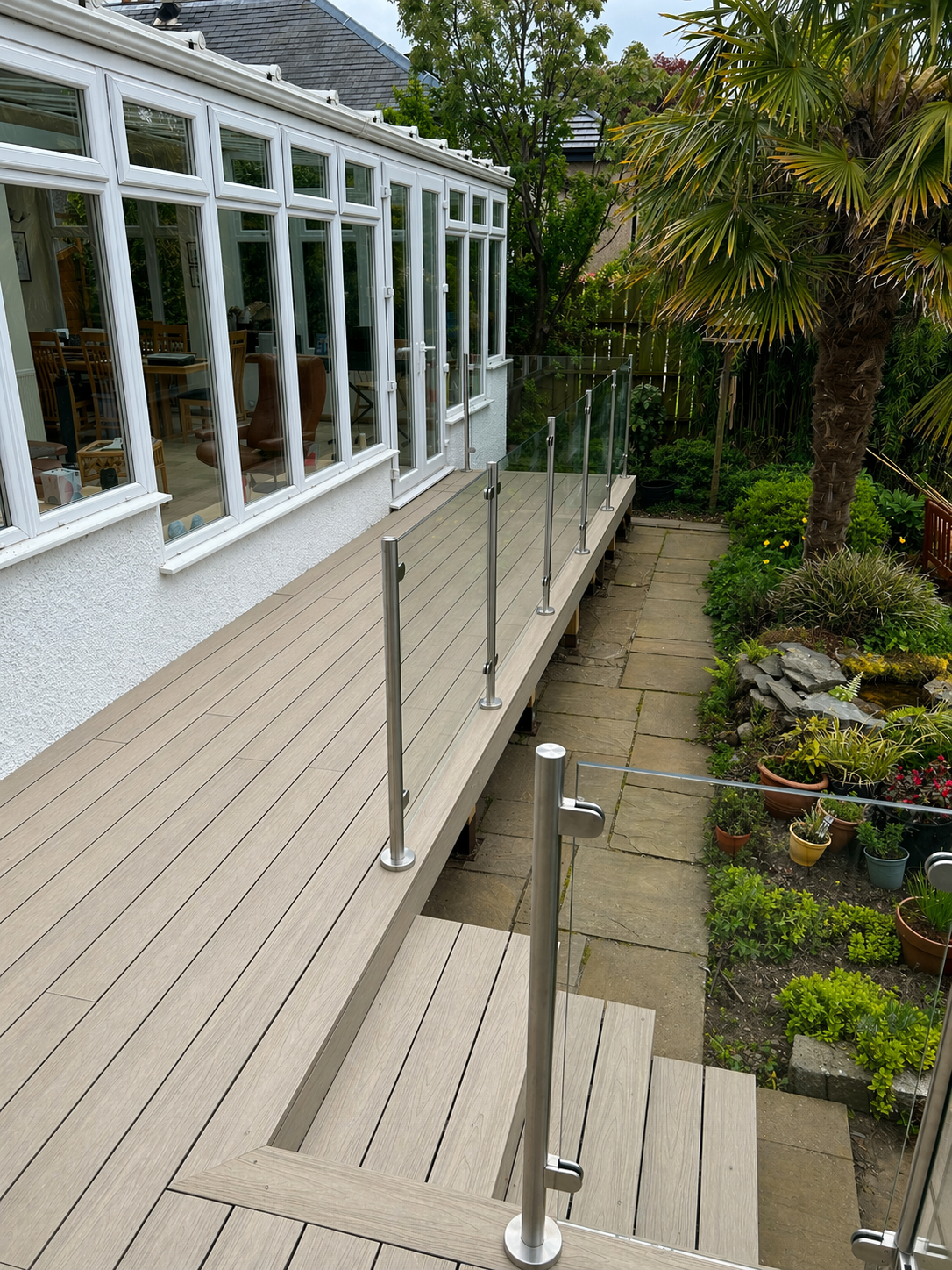 A backyard with a wooden deck and glass railing, next to a garden with plants, shrubs, and a palm tree, adjacent to a house with large windows.