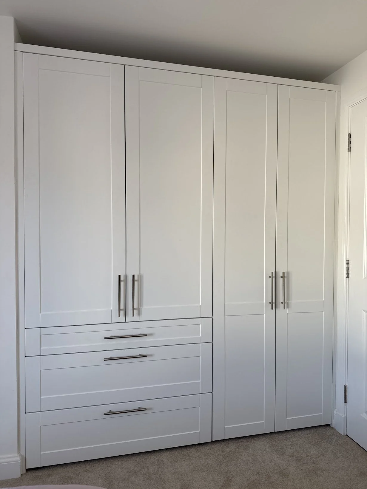 A white built-in closet with multiple doors and drawers, featuring silver handles, located in a room with beige carpeting and white walls.