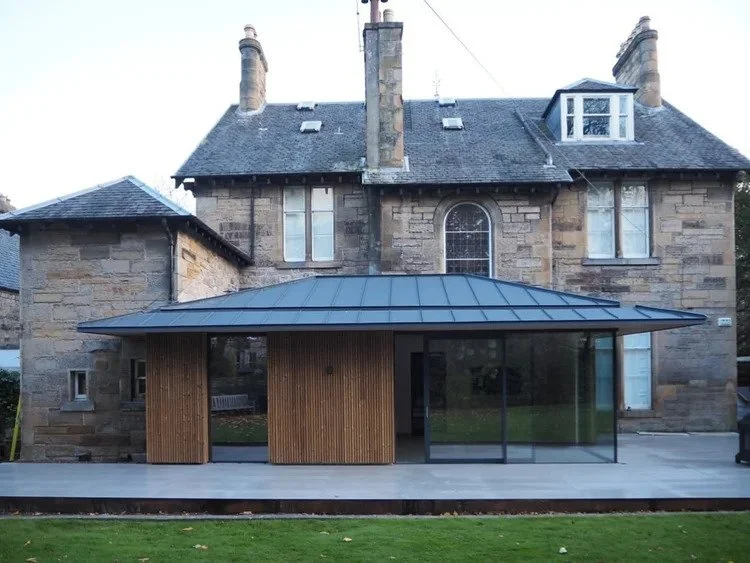 Modern glass and wood extension added to the back of a historic stone house, featuring a curved black metal roof and large sliding glass doors.