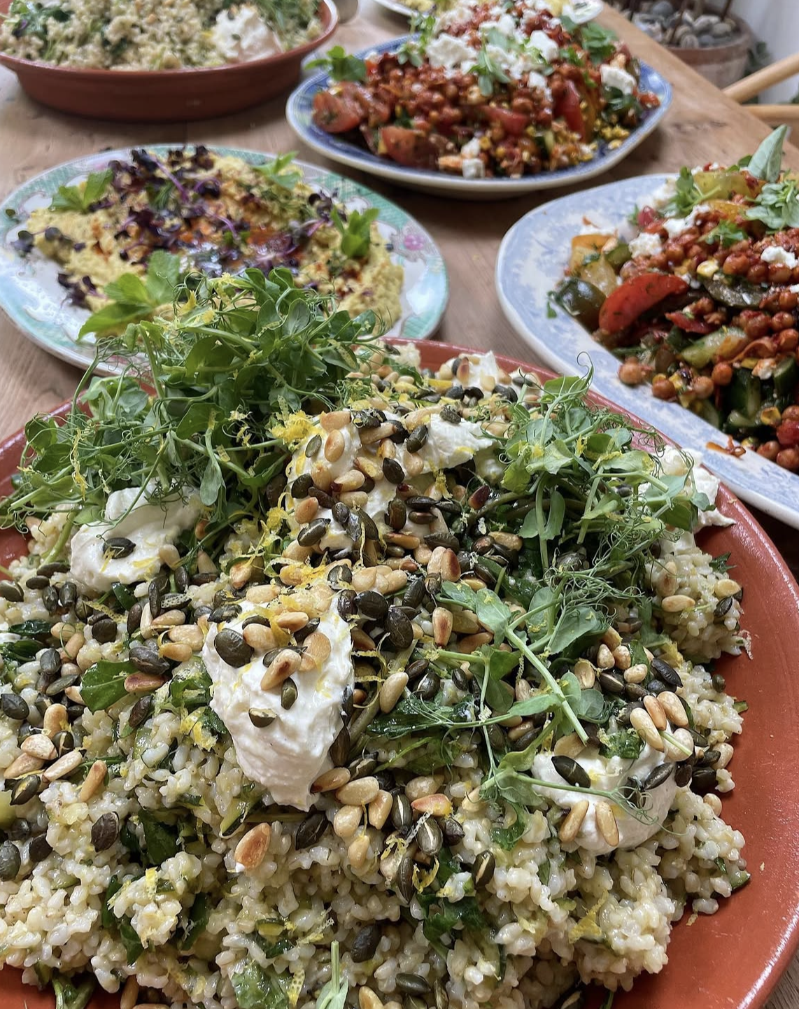 Close-up of a plate of grain salad topped with greens, seeds, and dollops of white creamy dressing, with several other colorful vegetable-based salads in the background.