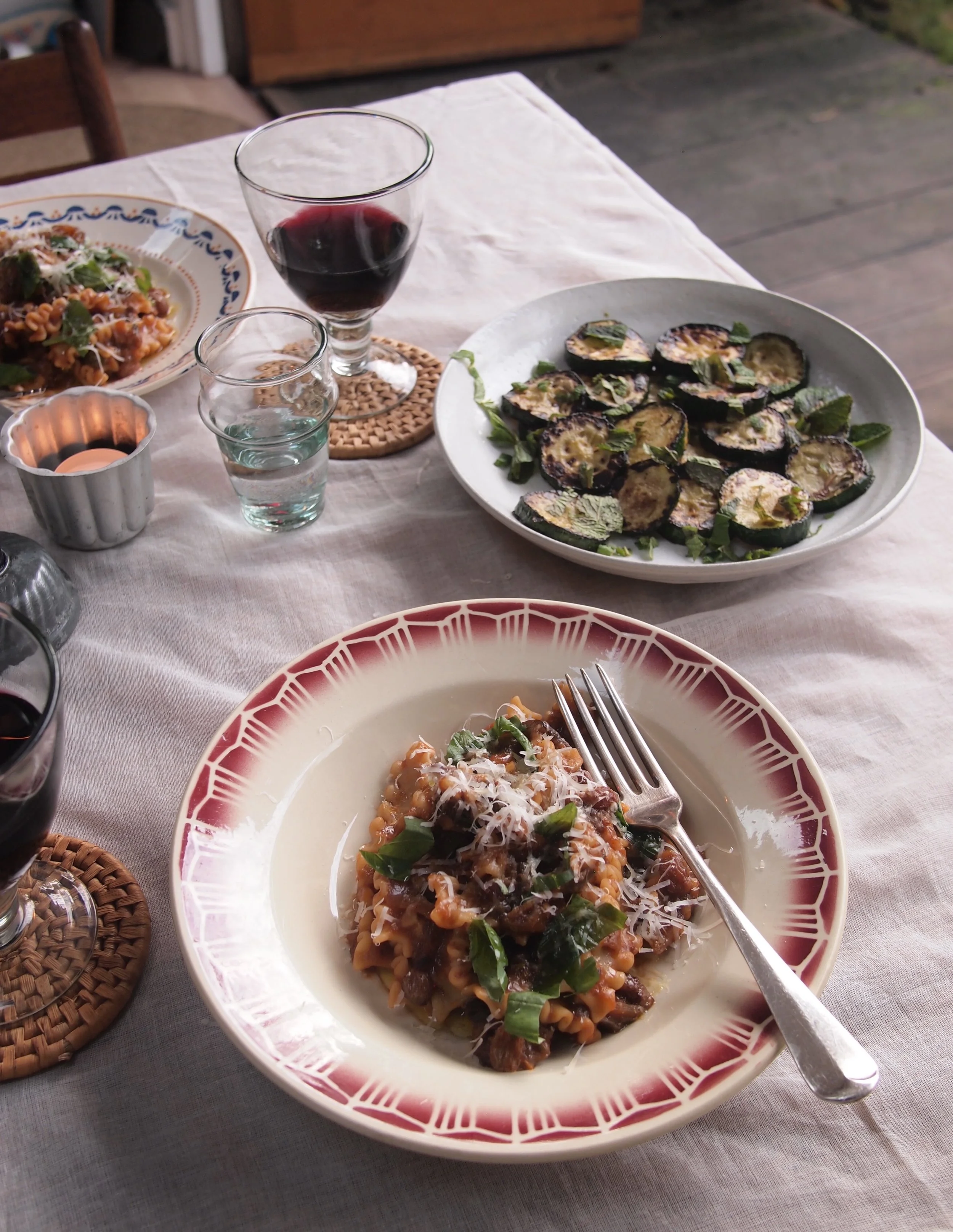 A dinner table set with plates of pasta, grilled zucchini slices, glasses of red wine, and water, on a white tablecloth, with a wooden chair and outdoor scenery in the background.
